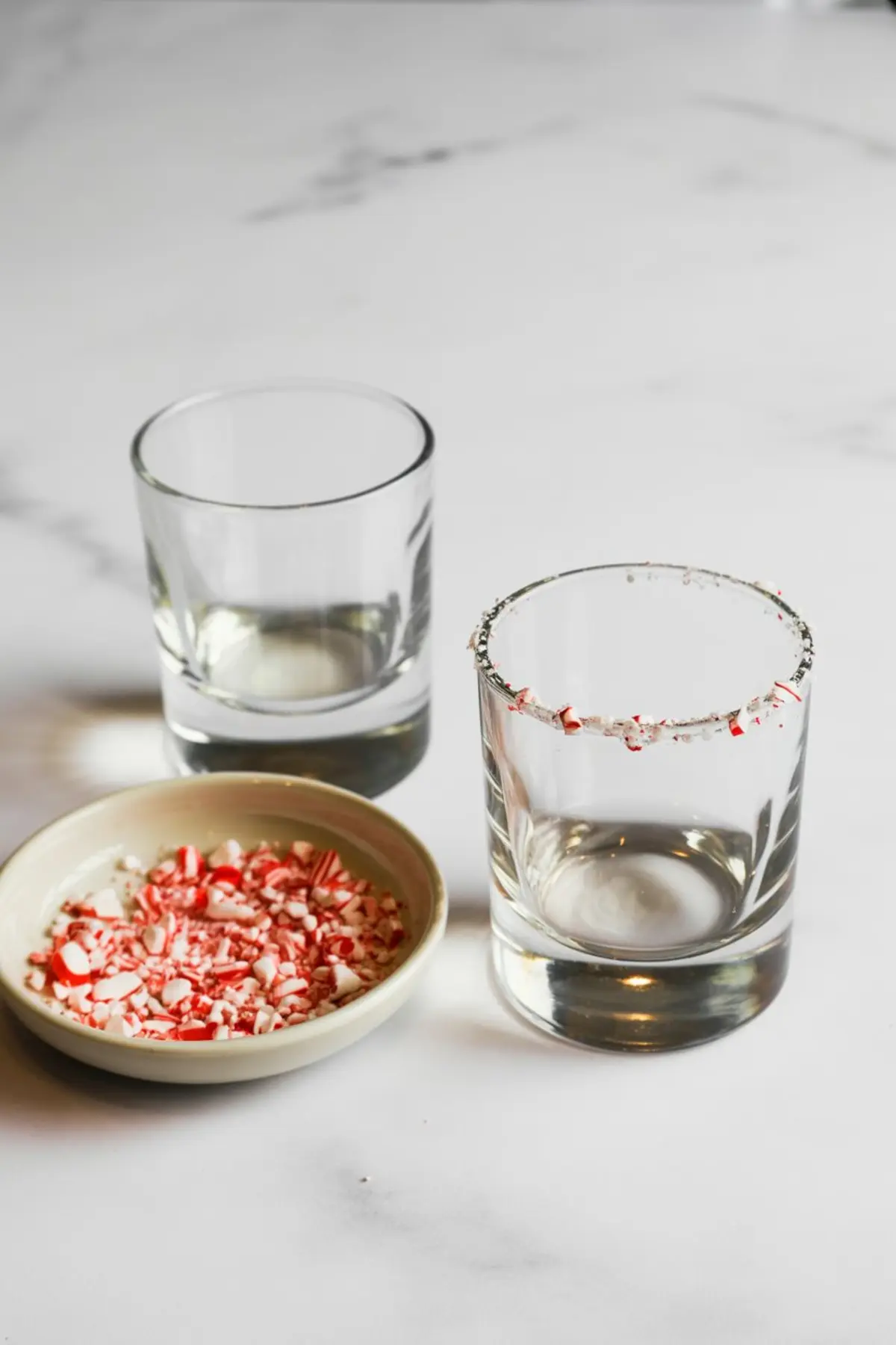 Two empty cocktail glasses on a white surface, one rimmed with crushed peppermint candy and a small dish of crushed peppermint beside them for garnishing holiday drinks.
