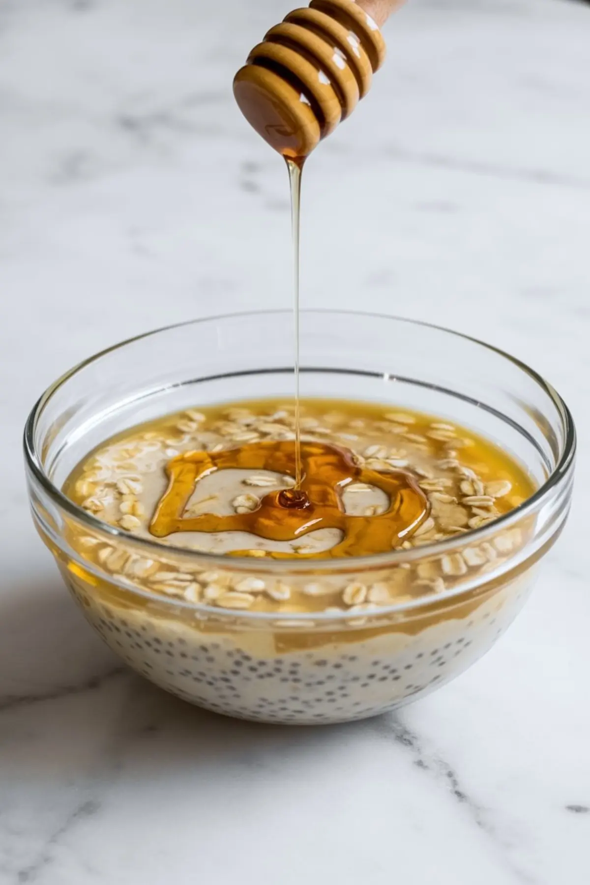 A glass bowl holds overnight oats with chia seeds and rolled oats as honey streams from a wooden dipper onto the surface.