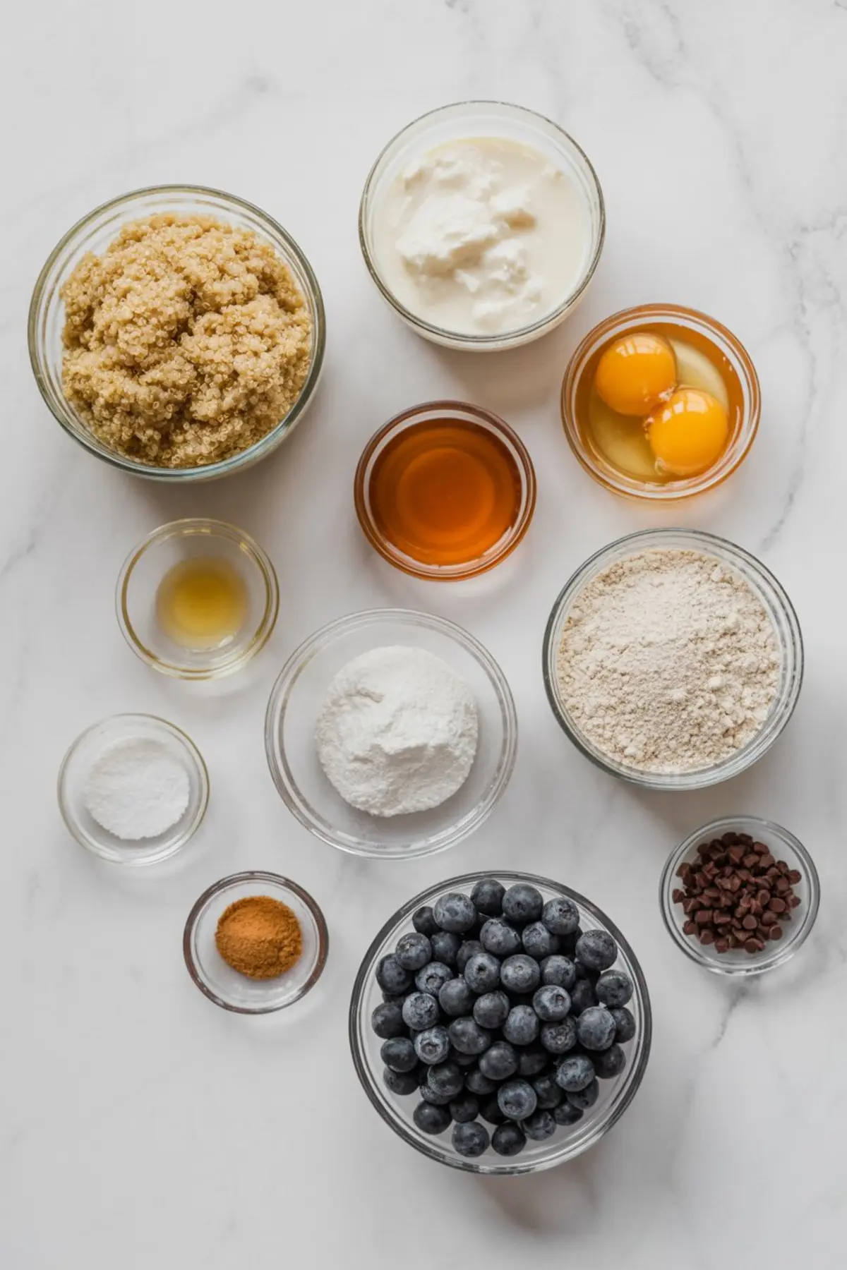 Flat lay of measured muffin ingredients in glass bowls including cooked quinoa, Greek yogurt, eggs, almond flour, maple syrup, baking powder, cinnamon, vanilla extract, blueberries, and mini chocolate chips on a marble background.
