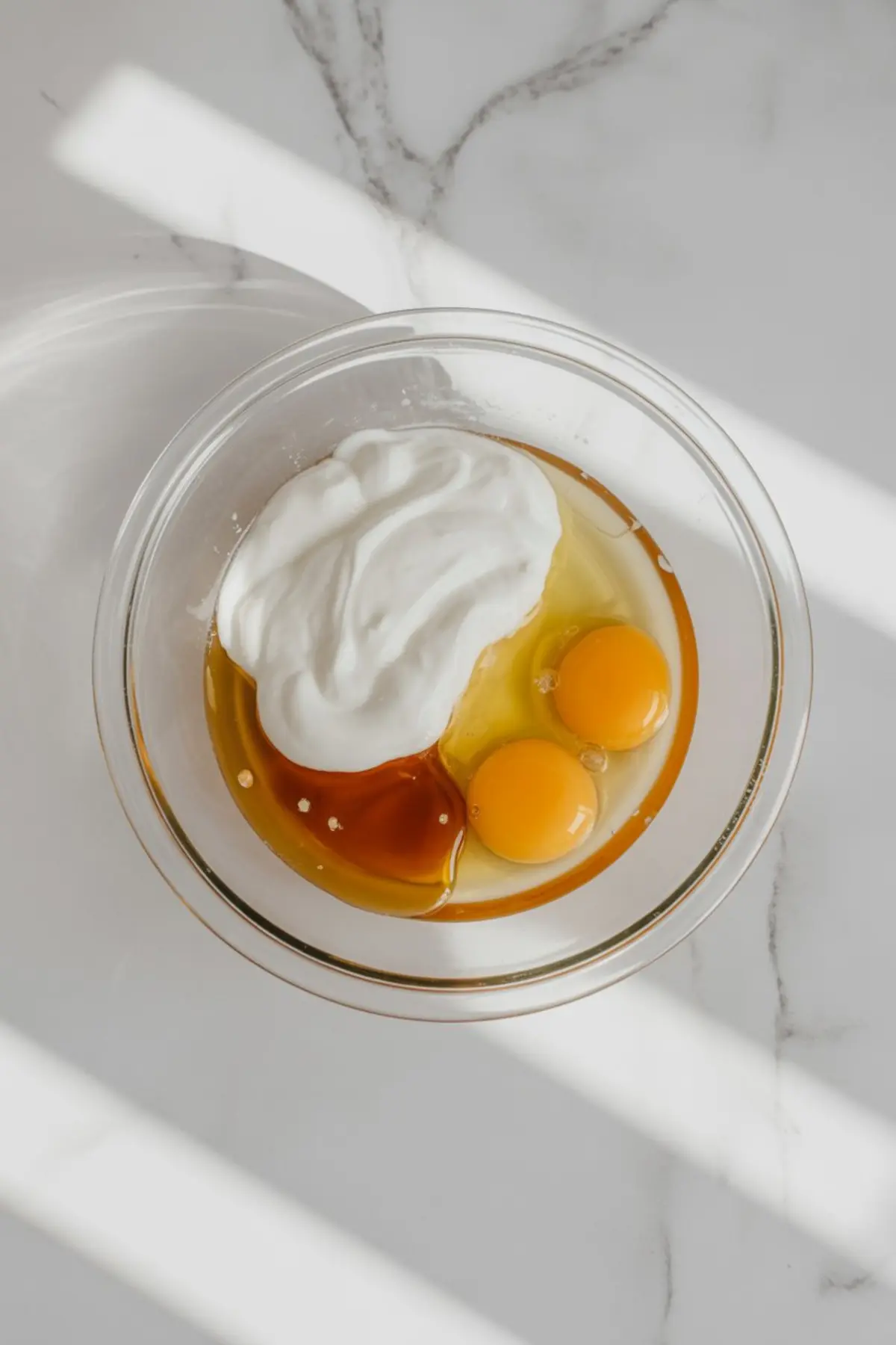 Top view of a glass bowl holding wet muffin ingredients including Greek yogurt, maple syrup, and two cracked eggs on a white marble surface with light shadows.