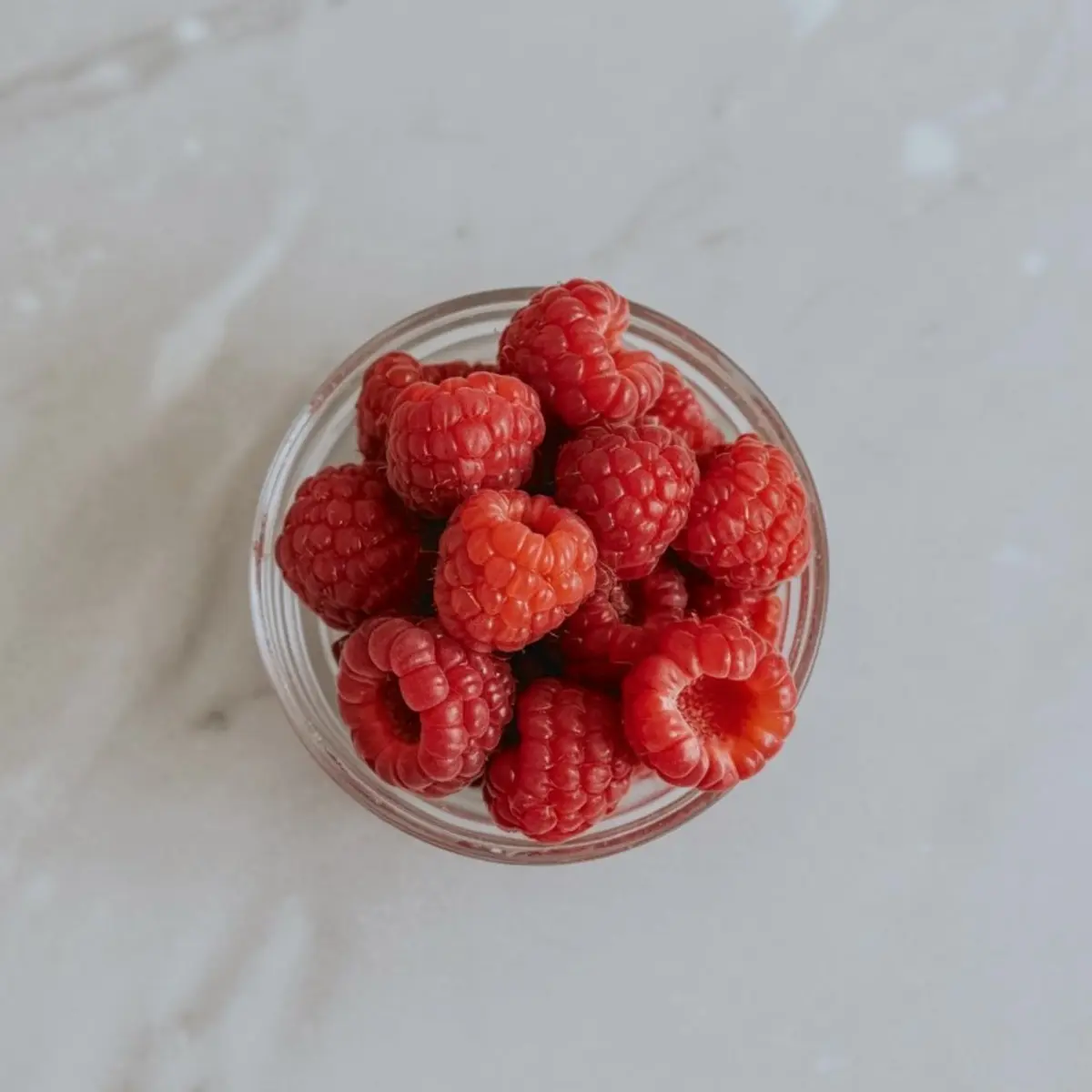 A glass bowl filled with fresh red raspberries on a light marble surface, showing vibrant berry texture and a clean minimal food styling look.
