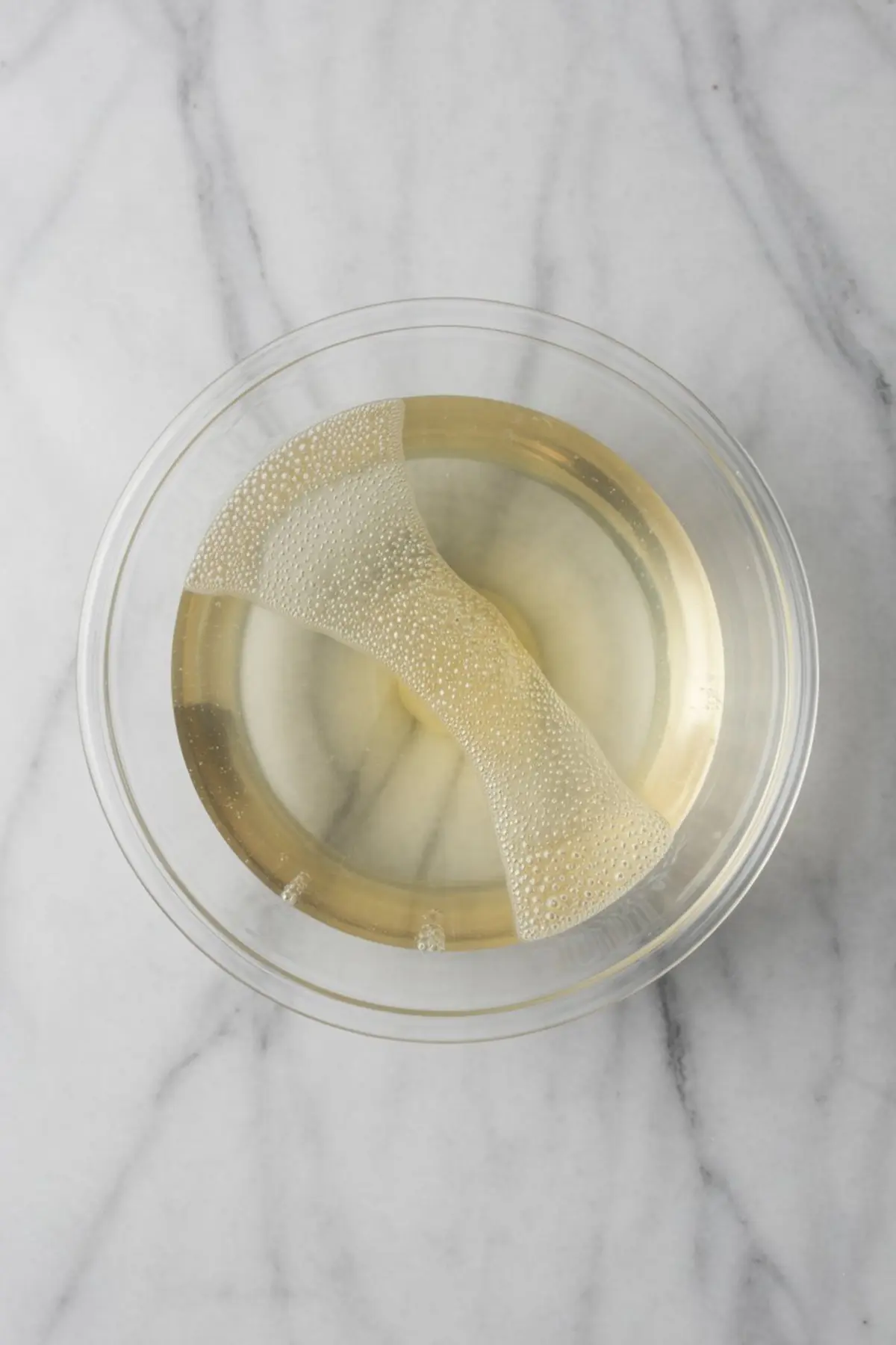 A glass bowl holding a hydrated gelatin sheet in pale golden liquid on a white marble surface, showing the preparation step for Prosecco jelly.
