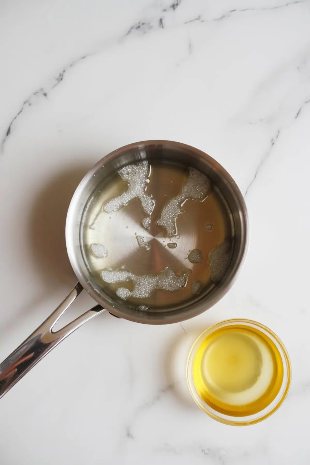 A stainless-steel saucepan with warm sugar mixture beside a small glass bowl of golden liquid on a white marble surface, showing a cooking step for Prosecco jelly preparation.
