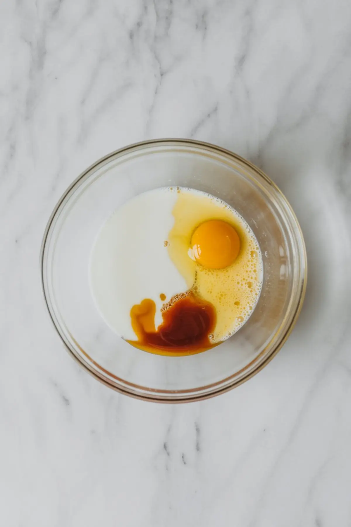 Clear bowl containing milk, an egg, and vanilla extract arranged separately before mixing, placed on a light marble surface.
