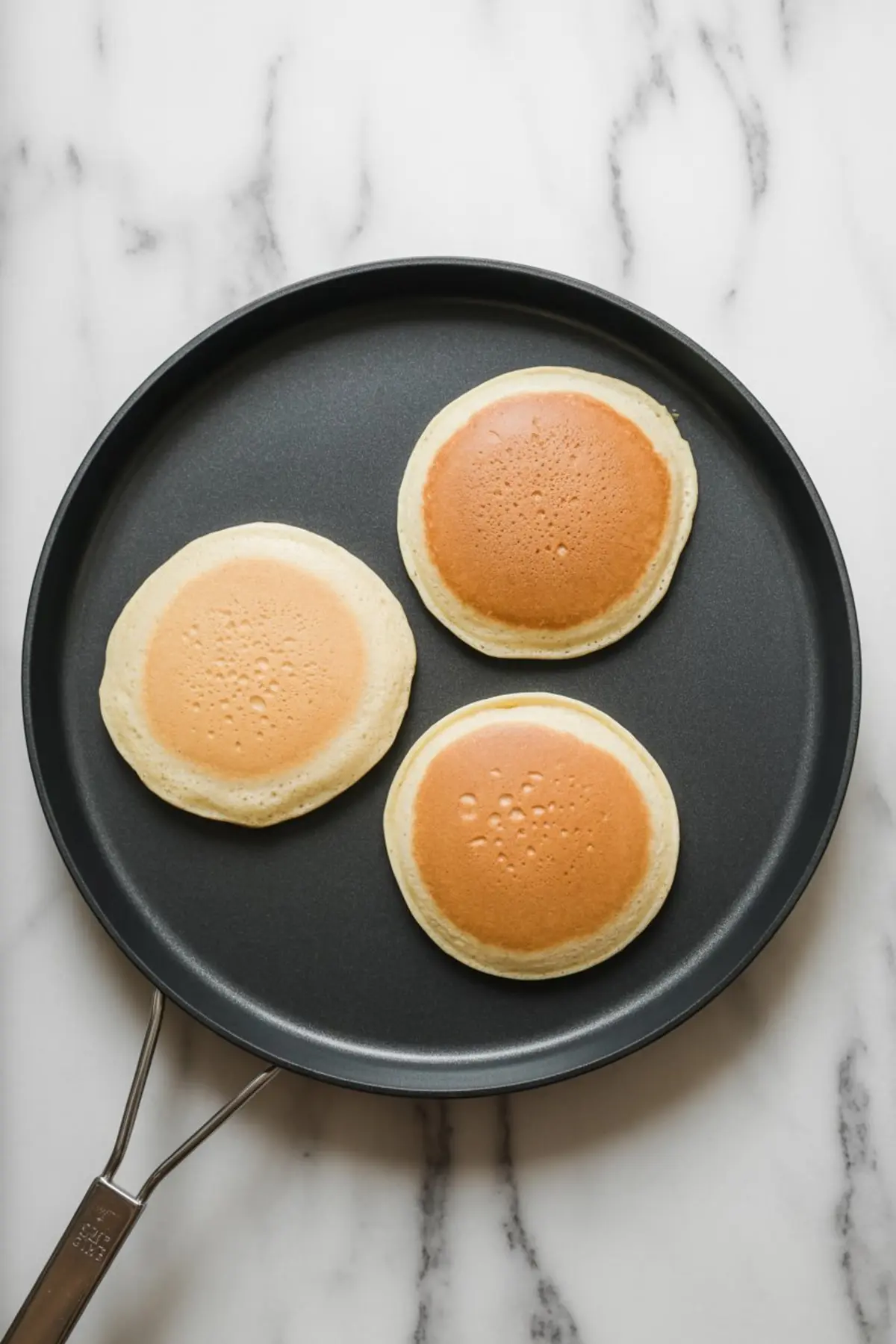 Three golden-brown pancakes cooking on a non-stick skillet over a marble countertop, showing smooth texture and even browning.
