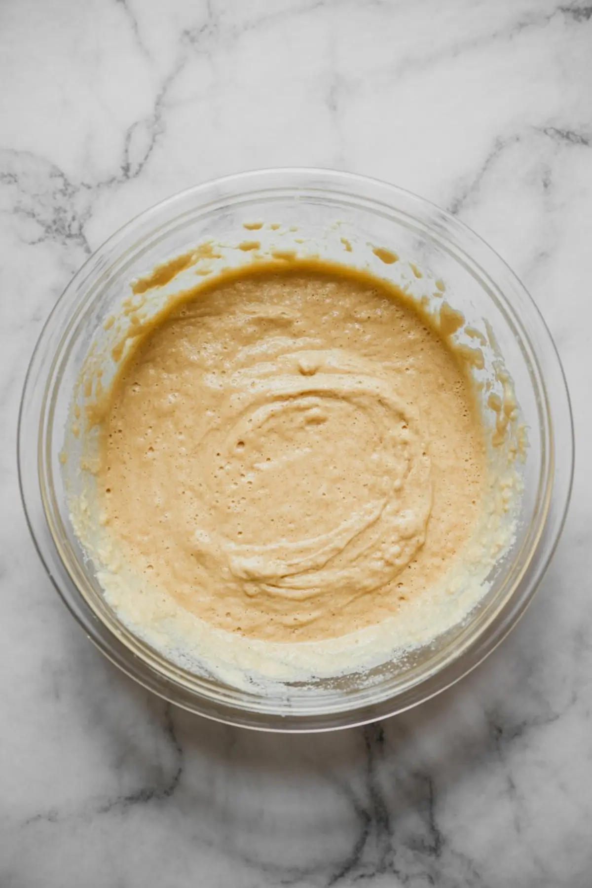 Bowl of freshly mixed pancake batter with a thick, smooth consistency in a glass bowl on a white marble surface.
