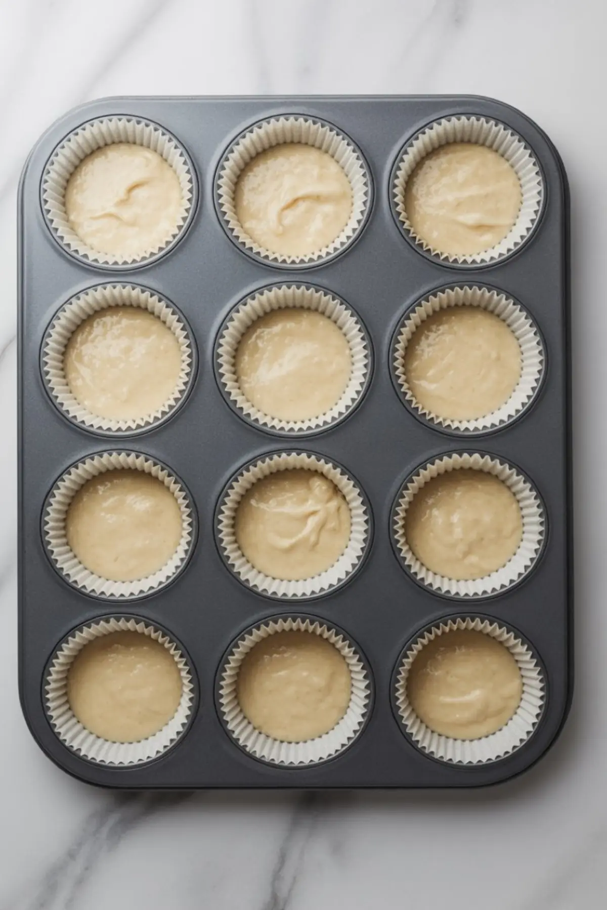 Muffin tin filled with cupcake batter in white liners, arranged on a marble surface and ready to be baked for classic vanilla cupcakes.
