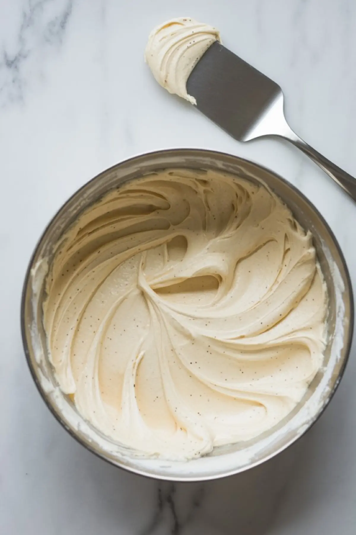 Bowl of whipped vanilla bean buttercream frosting with visible specks, accompanied by a metal spatula coated with a smooth swipe of the creamy frosting.
