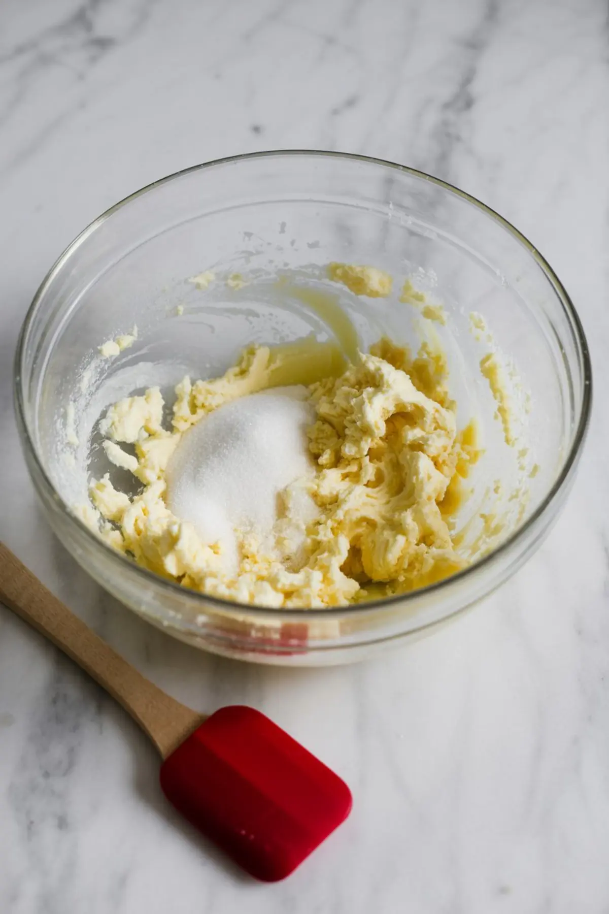 Glass mixing bowl with creamed butter and sugar mixture on a marble counter, next to a red spatula, capturing the base for homemade vanilla cupcake batter.
