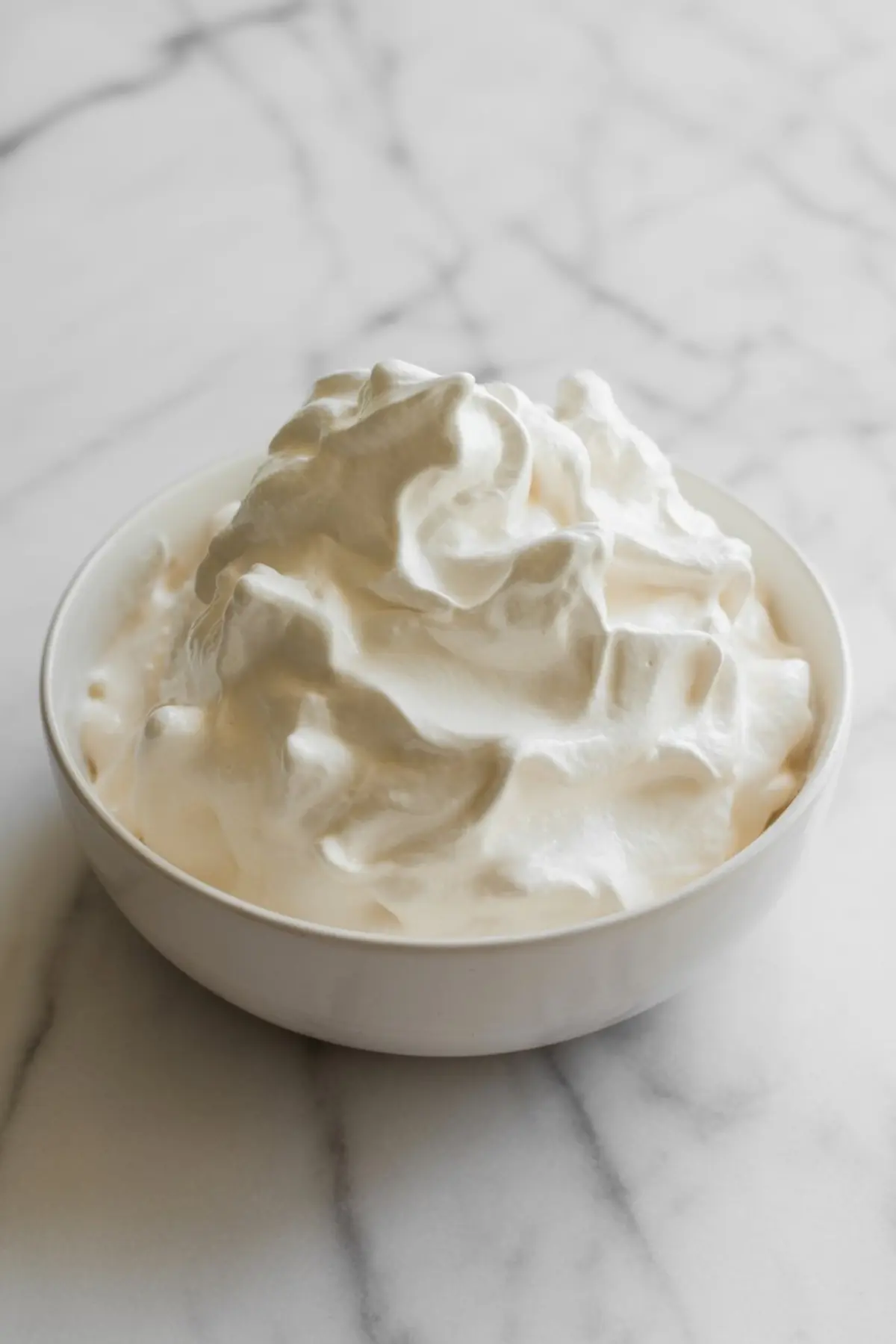 Fluffy whipped cream piled high in a white bowl on a light marble surface, ideal for topping desserts or folding into mousse recipes.
