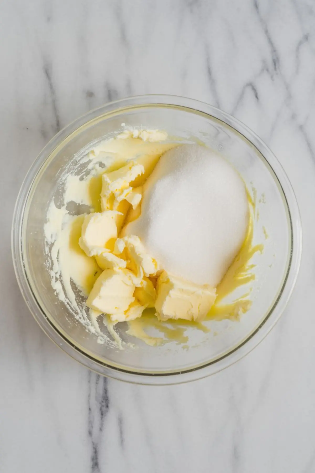 Softened butter and granulated sugar in a glass mixing bowl on a marble countertop, ready to be creamed for sugar cookie batter.