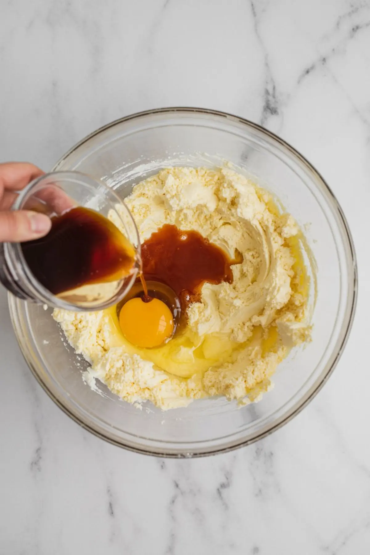 Pouring vanilla extract into a glass mixing bowl filled with softened butter, sugar, and a cracked egg for sugar cookie dough.