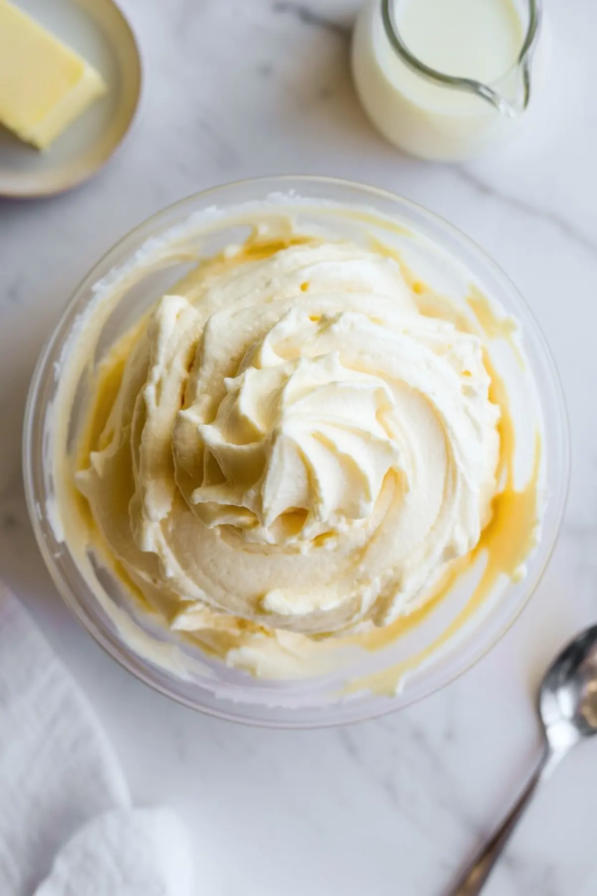 Fluffy vanilla buttercream frosting swirled in a glass bowl with visible hints of creamy yellow and soft white texture.