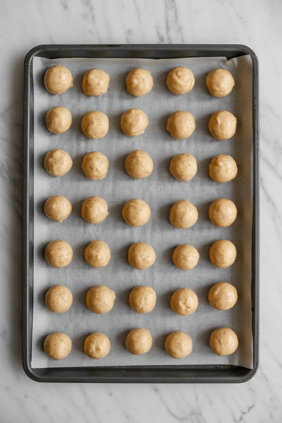 Unbaked vanilla cookie dough balls lined up on a parchment-lined baking sheet, evenly spaced and ready to go into the oven.
