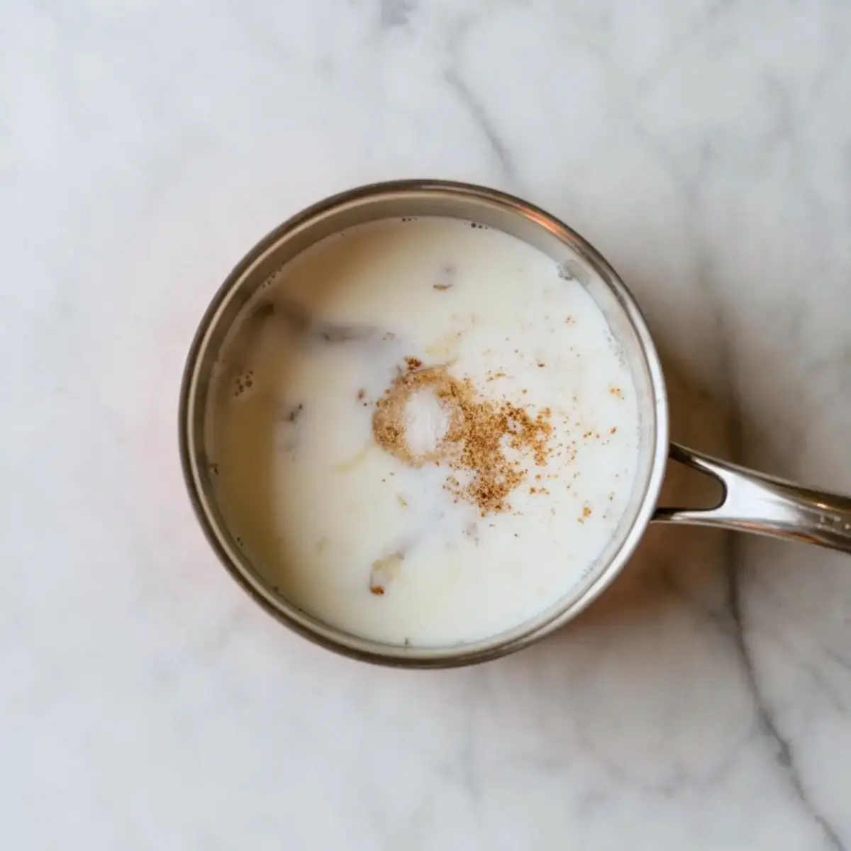 Top view of a stainless steel saucepan filled with milk, spices, and visible chunks of clove and cinnamon, resting on a marble countertop, showing the beginning stage of homemade eggnog preparation.