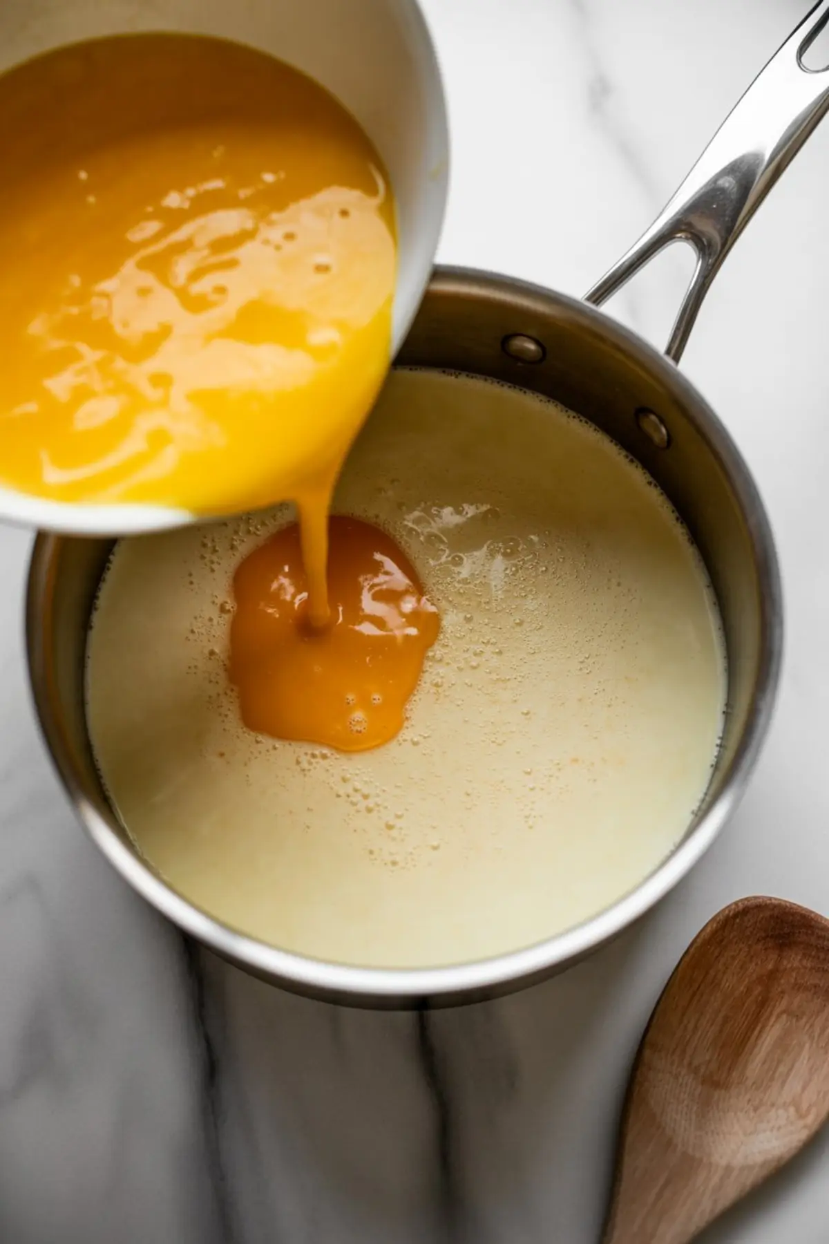 Bright kitchen scene of golden egg yolks being poured from a white bowl into a saucepan of warm milk mixture on a marble surface, capturing the blending step for homemade eggnog.