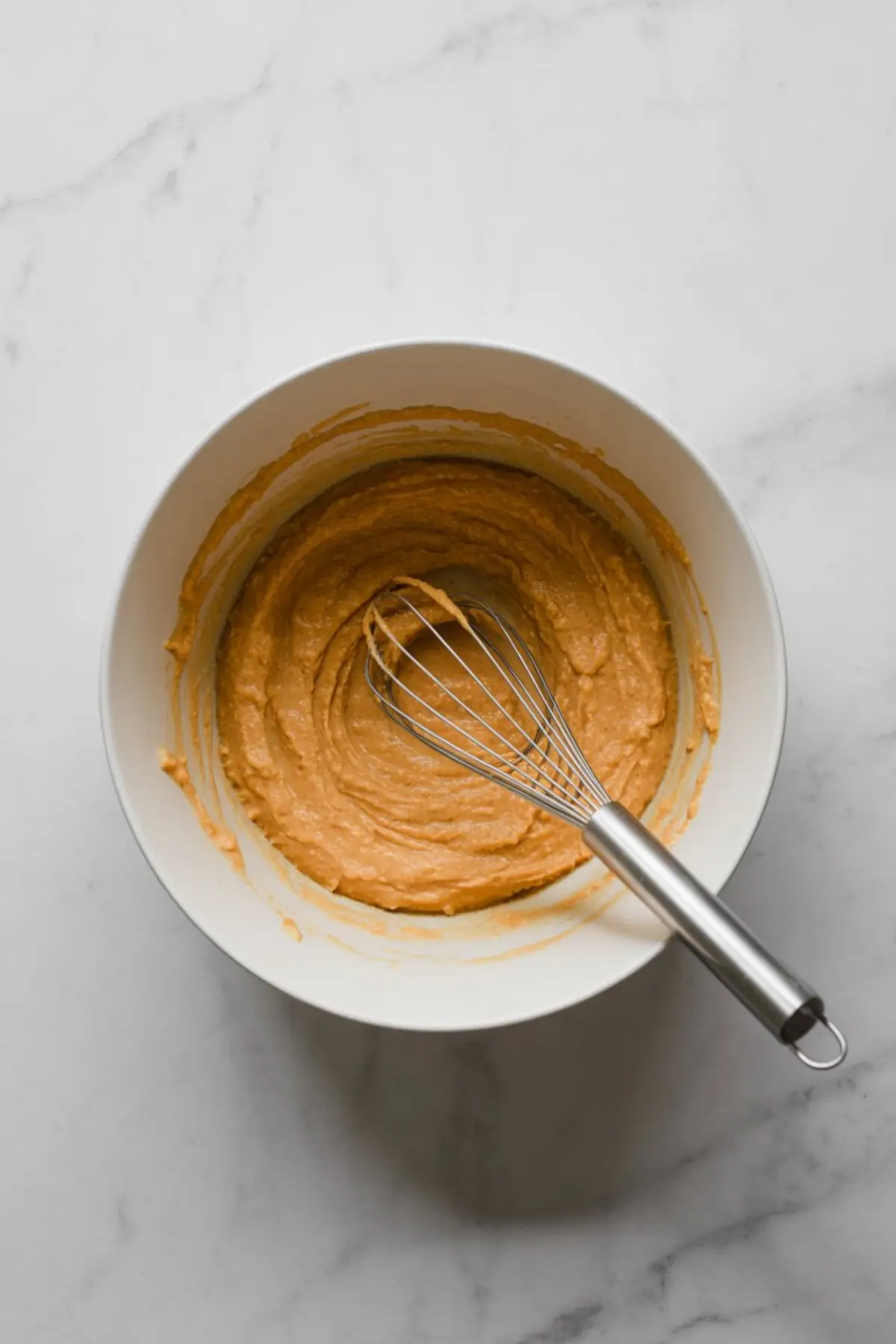 Smooth orange sweet potato cake batter in a white mixing bowl with a metal whisk on a white marble background, ready to be poured into a baking dish for dessert preparation.
