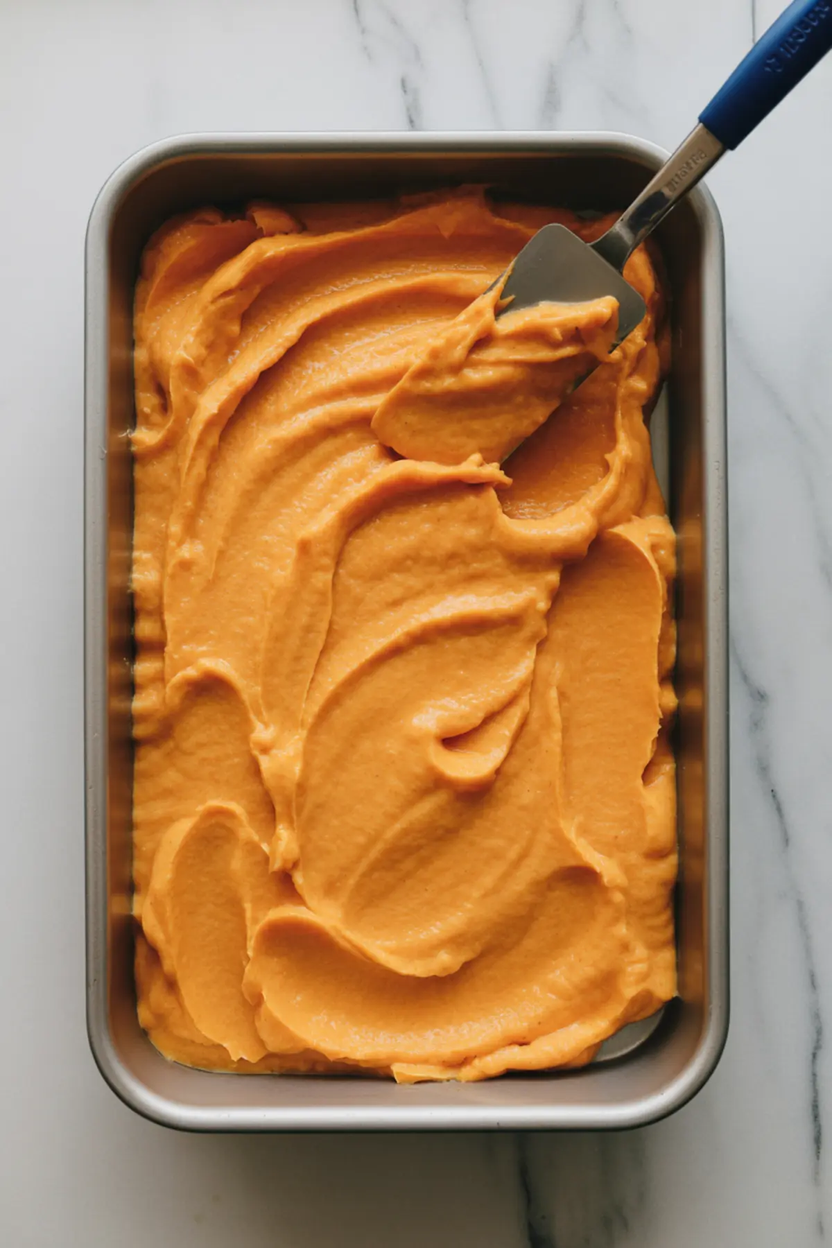 Creamy sweet potato batter evenly spread in a metal baking pan with a spatula, ready for the crumb topping and oven baking.
