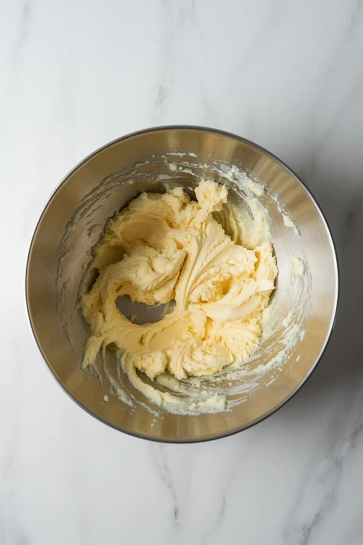 Metal mixing bowl filled with whipped butter and sugar mixture, showing the creamy, pale yellow texture of the first step in making cookie dough.