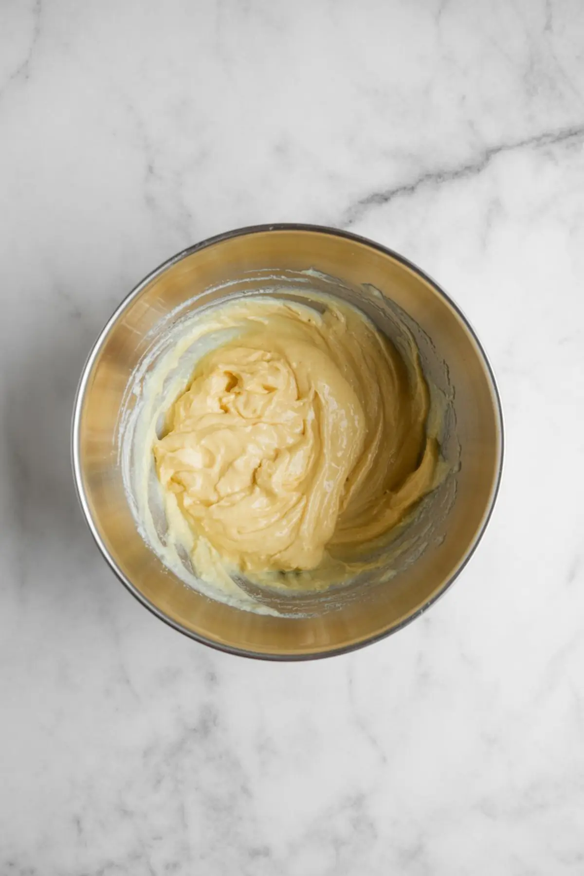 Top-down view of a mixing bowl with thick, creamy tiramisu cookie batter in pale yellow, resting on a marble counter.