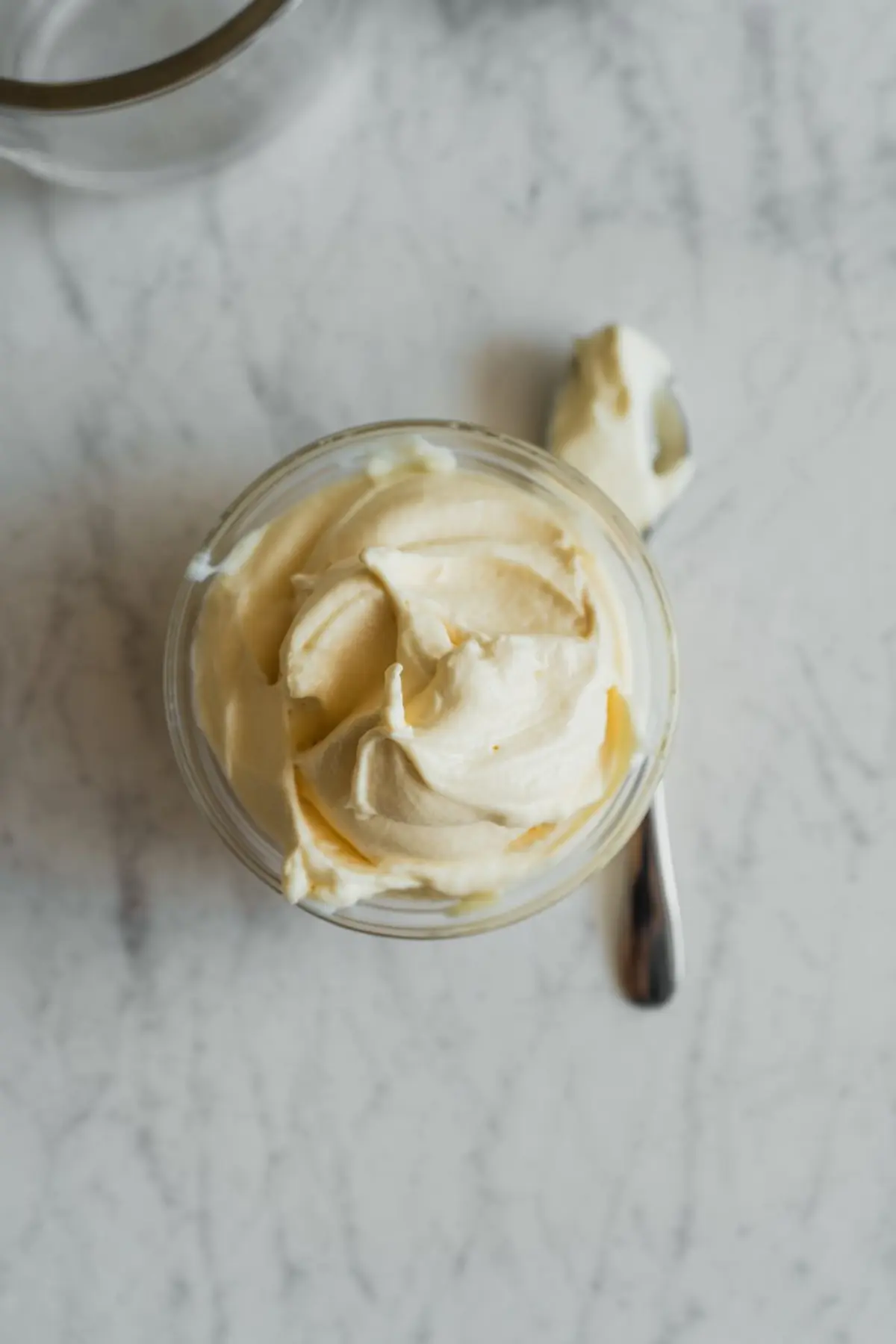 Glass jar of smooth mascarpone cream filling with a spoon beside it, photographed from above on a light marble counter.