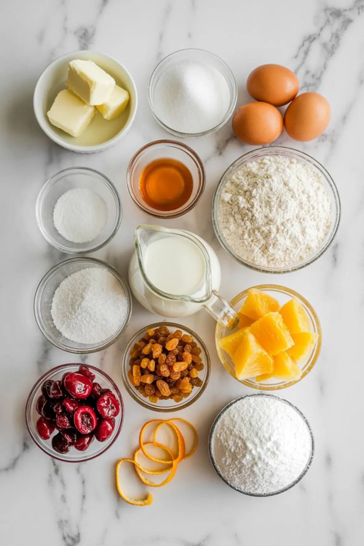 Glass bowls and measuring cups filled with ingredients for vintage Christmas cake, including butter, sugar, eggs, flour, milk, vanilla extract, salt, baking powder, dried cherries, golden raisins, orange segments, and powdered sugar, all arranged on a white marble background.