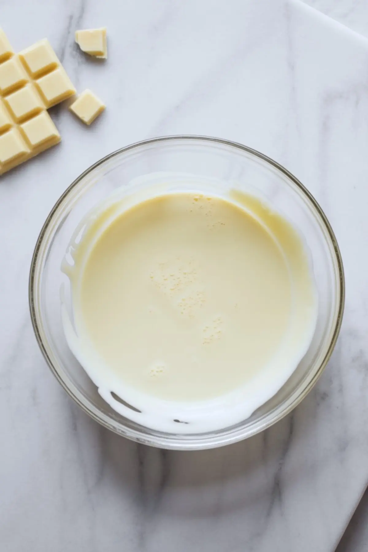 Melted white chocolate in a glass bowl on a white marble surface, with small chunks of white chocolate bar placed beside the bowl.