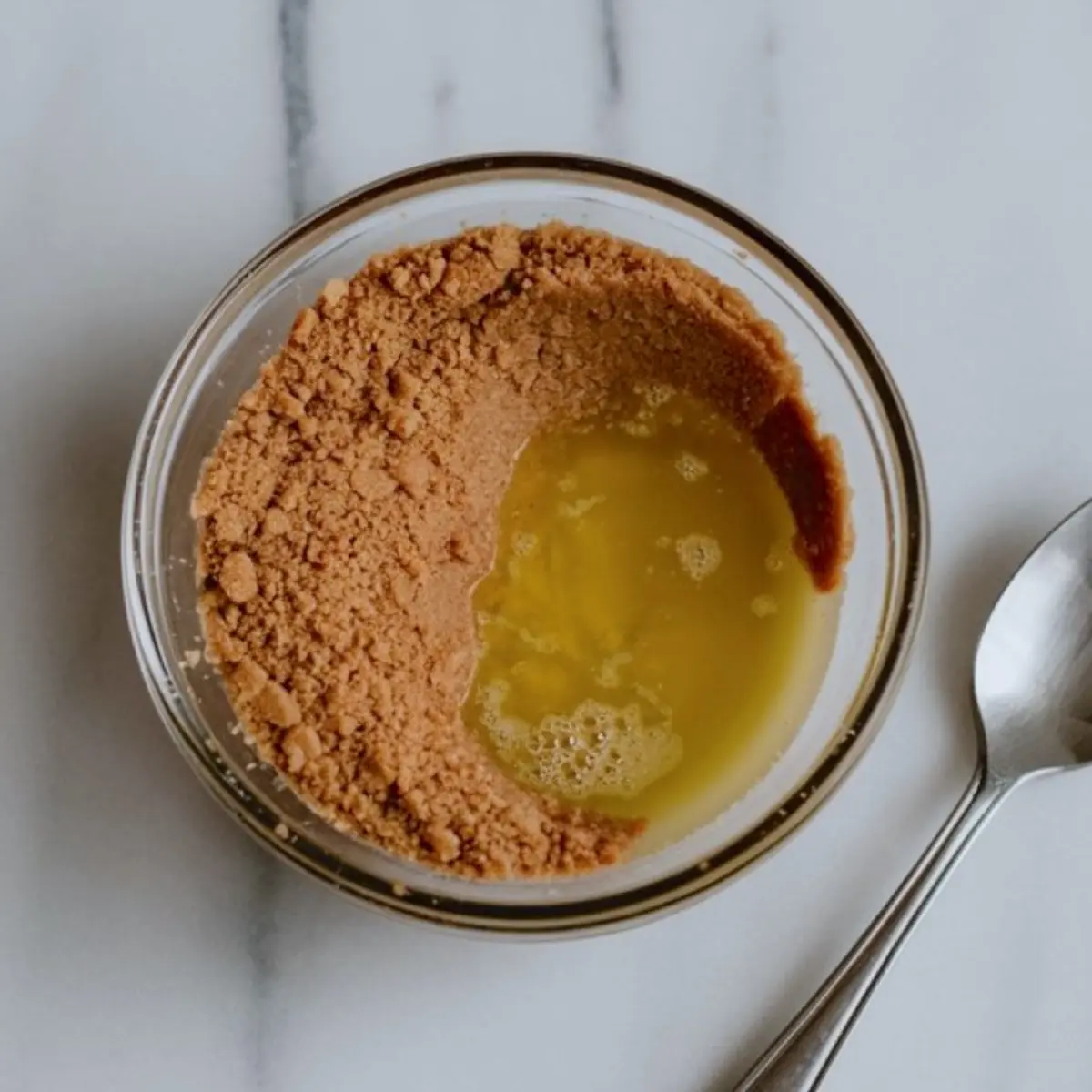 Graham cracker crumbs partially mixed with melted butter in a small glass bowl on a white surface, with a metal spoon placed next to it.