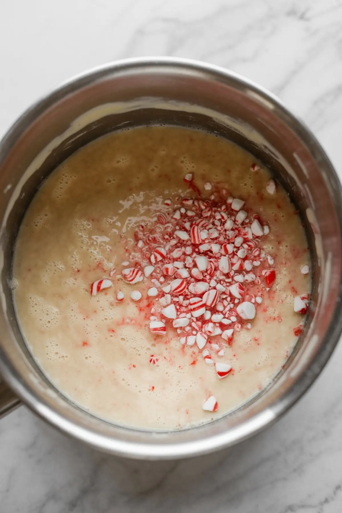 Top-down view of a metal mixing bowl filled with pale yellow fudge batter, topped with crushed peppermint candy pieces, sitting on a white marble surface.
