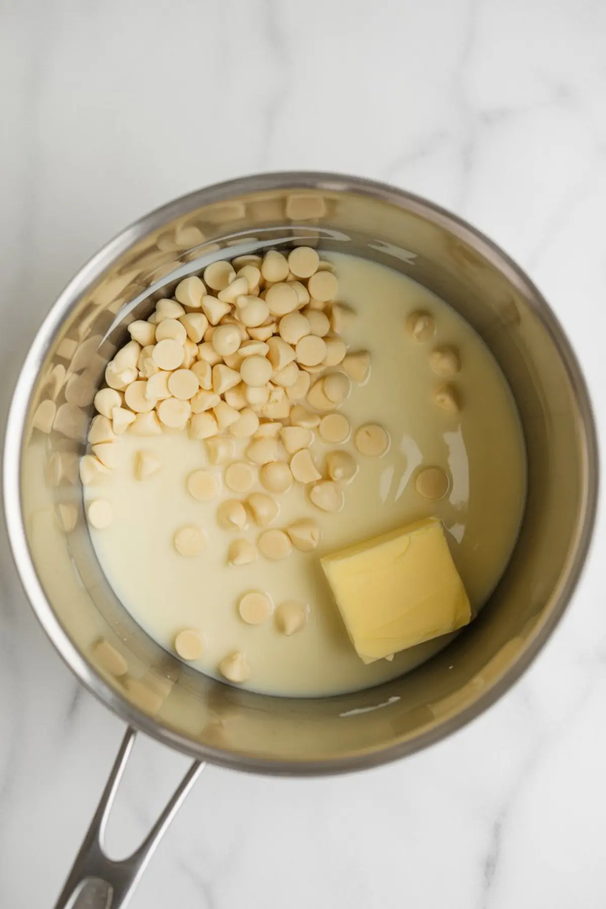 Unmelted white chocolate chips, a block of butter, and sweetened condensed milk in a saucepan, ready to be heated for a fudge recipe.
