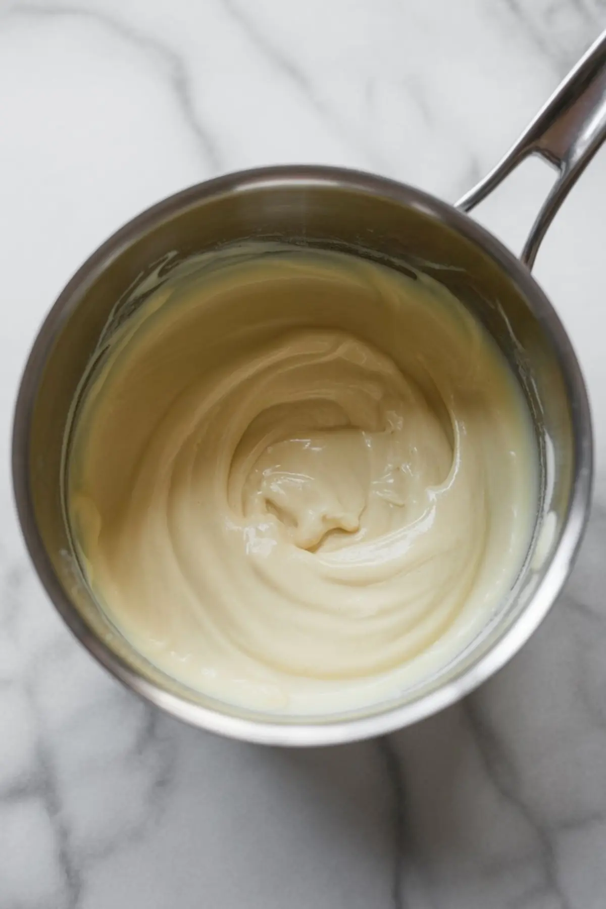 Smooth, melted white chocolate mixture in a saucepan with a creamy consistency, set on a light marble countertop.
