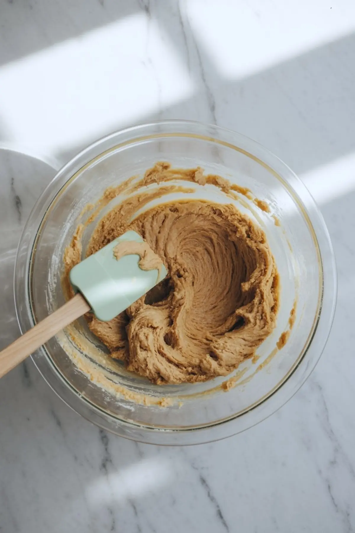 Creamy almond flour cookie dough swirled in a glass mixing bowl with a light green silicone spatula, placed on a sunlit marble countertop.