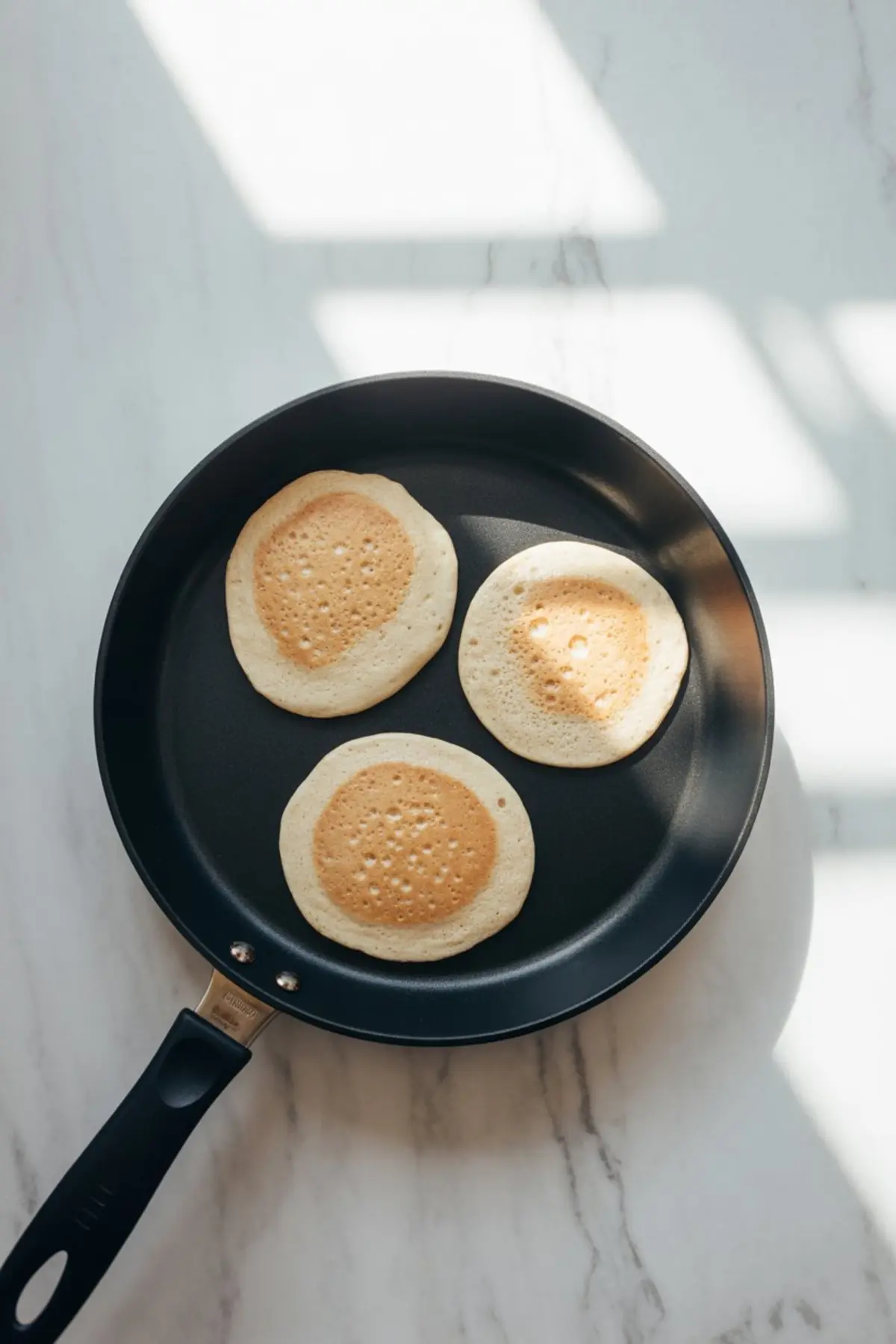 Three almond flour pancakes cooking in a nonstick skillet with golden centers and light edges on a marble stovetop background.

