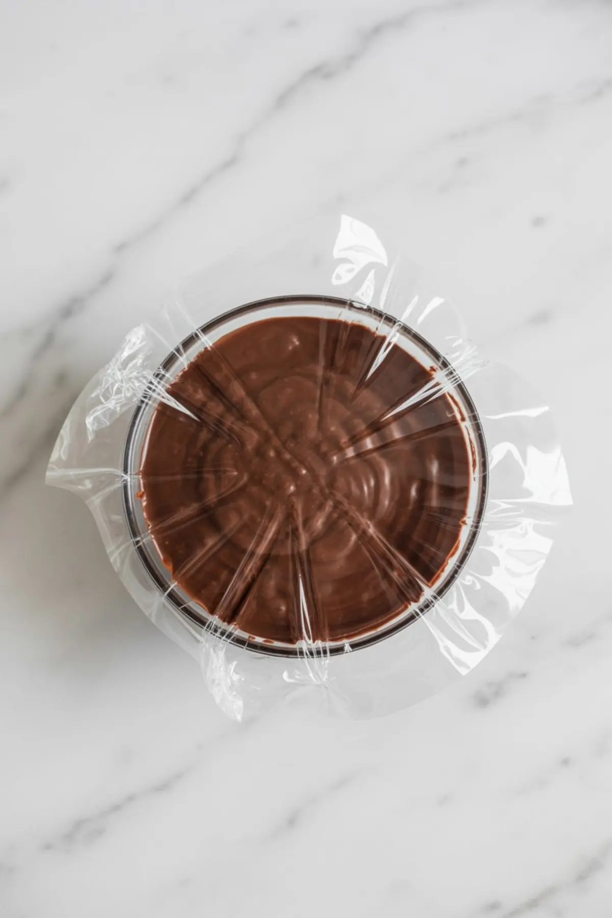 Chocolate avocado truffle mixture rests in a round container sealed with plastic wrap on a marble surface, showing a smooth chilled vegan chocolate base.
