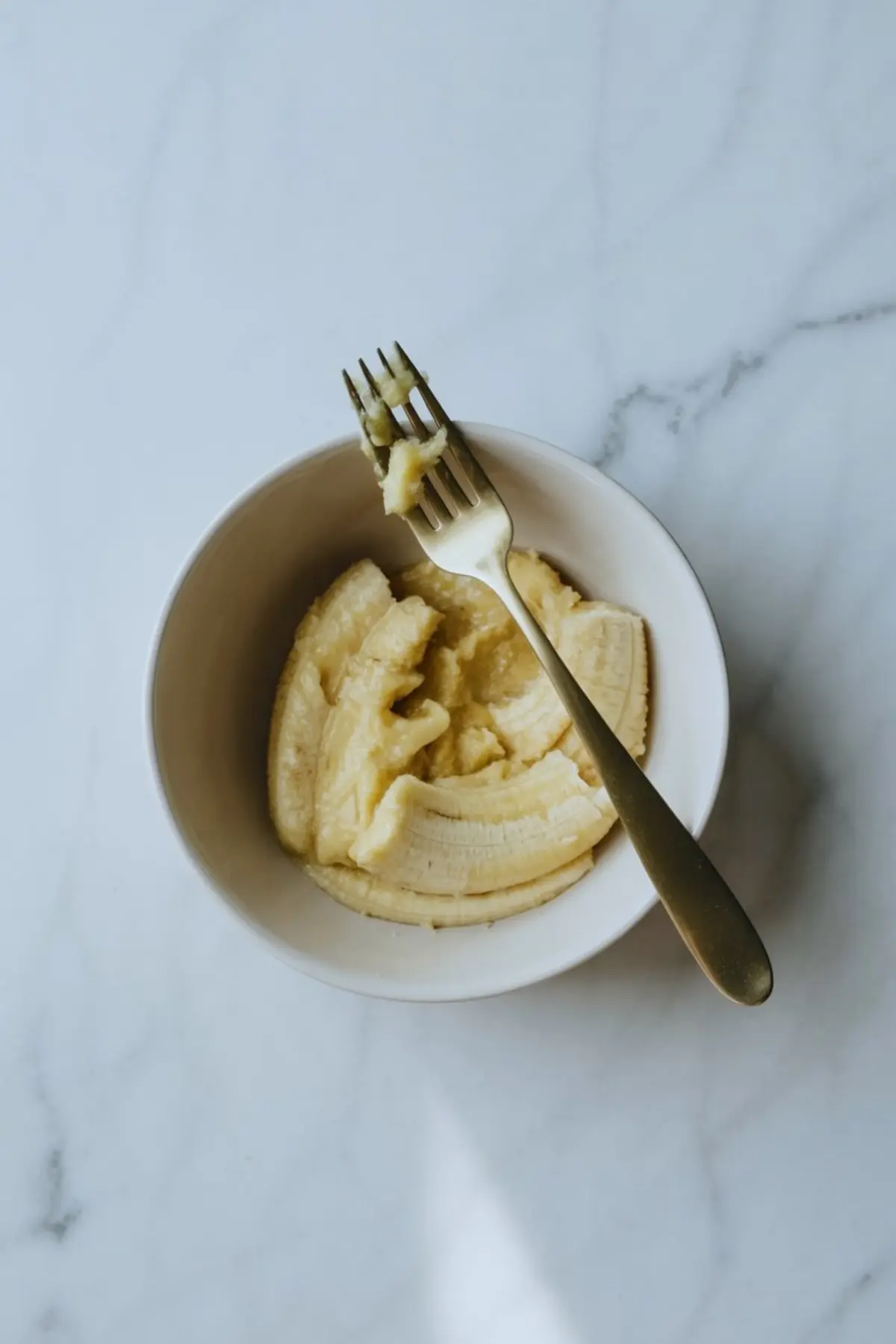 White bowl with partially mashed bananas and a fork resting inside, placed on a marble counter.
