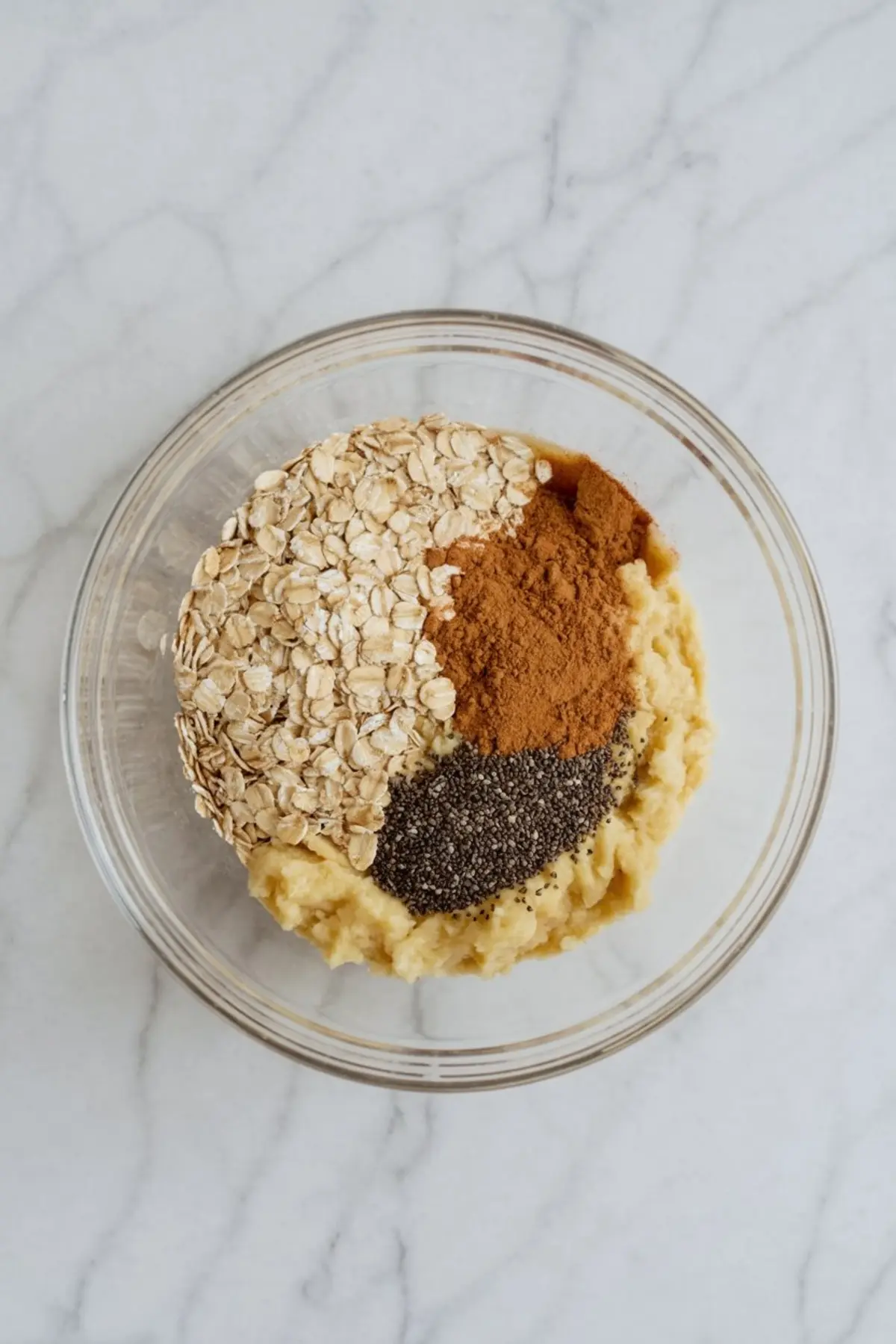 Clear glass bowl containing rolled oats, mashed banana, cinnamon, and chia seeds arranged in separate sections before mixing.
