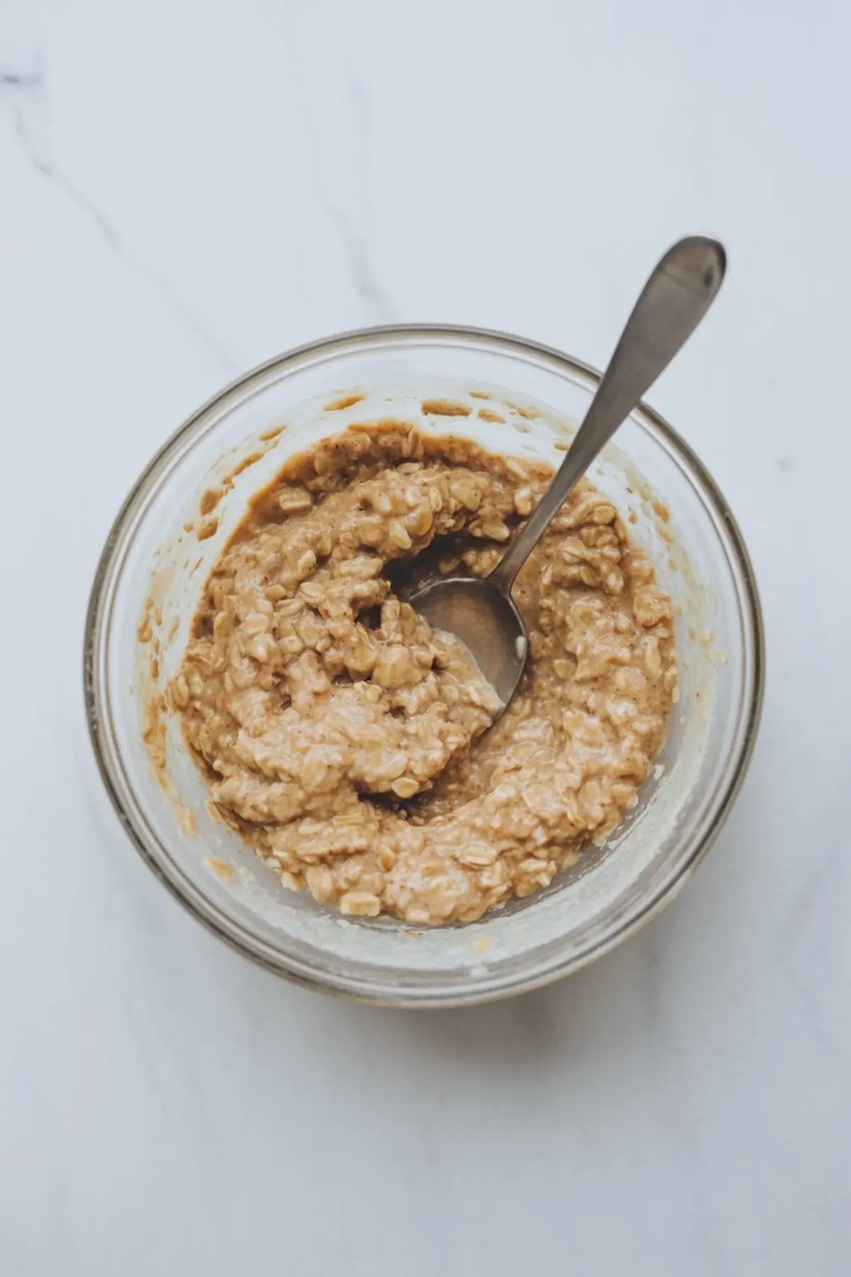 Thick banana oat mixture stirred together in a glass bowl with a metal spoon, showing a creamy and well-combined consistency.
