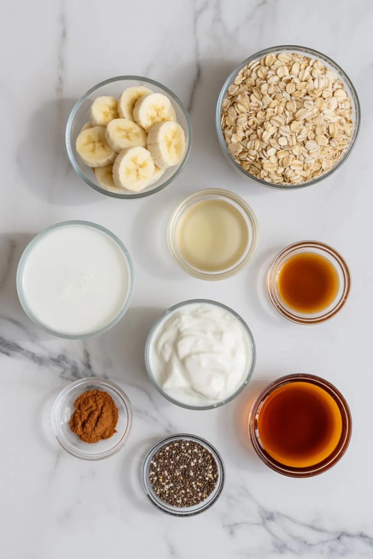 Flat lay of ingredients for banana overnight oats including rolled oats, banana slices, milk, yogurt, chia seeds, cinnamon, maple syrup, and vanilla extract arranged in glass bowls on a white marble surface.
