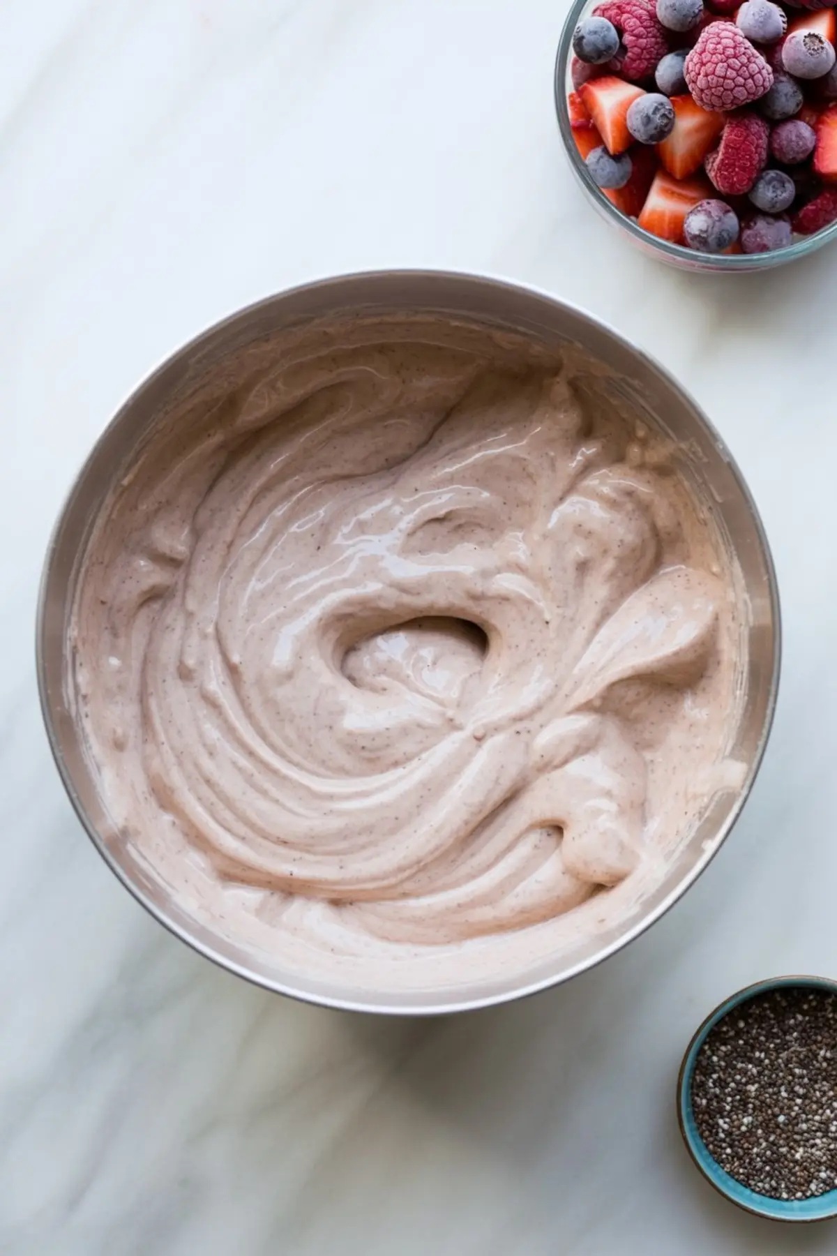 Bowl of thick, blended yogurt protein mixture beside a glass bowl of frozen mixed berries and a small bowl of chia seeds, set on a white marble surface.
