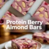 Close-up of sliced protein berry almond bars topped with whole almonds and mixed berries, featuring a creamy pink protein layer over a chewy nut base on a marble background.