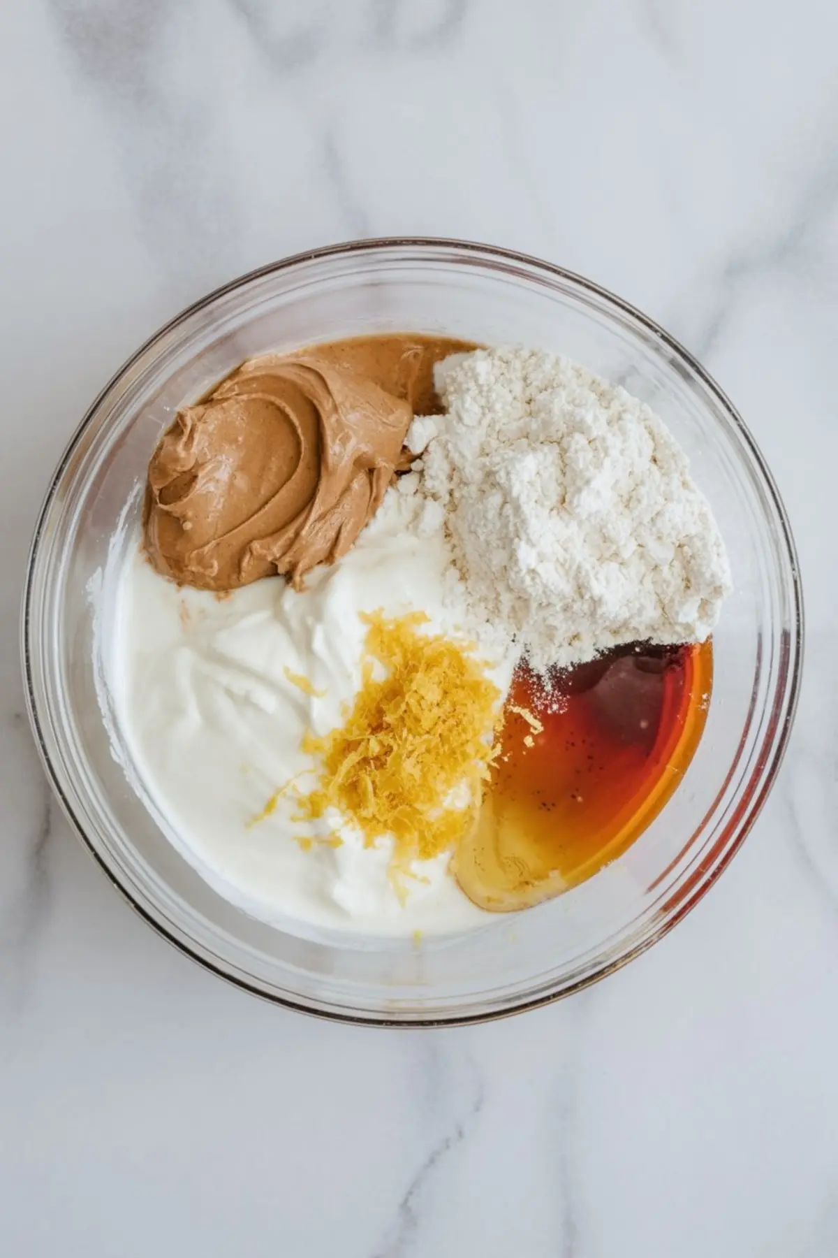 Glass bowl with measured ingredients including Greek yogurt, peanut butter, protein powder, lemon zest, and maple syrup, arranged neatly for a no-bake protein dessert recipe.

