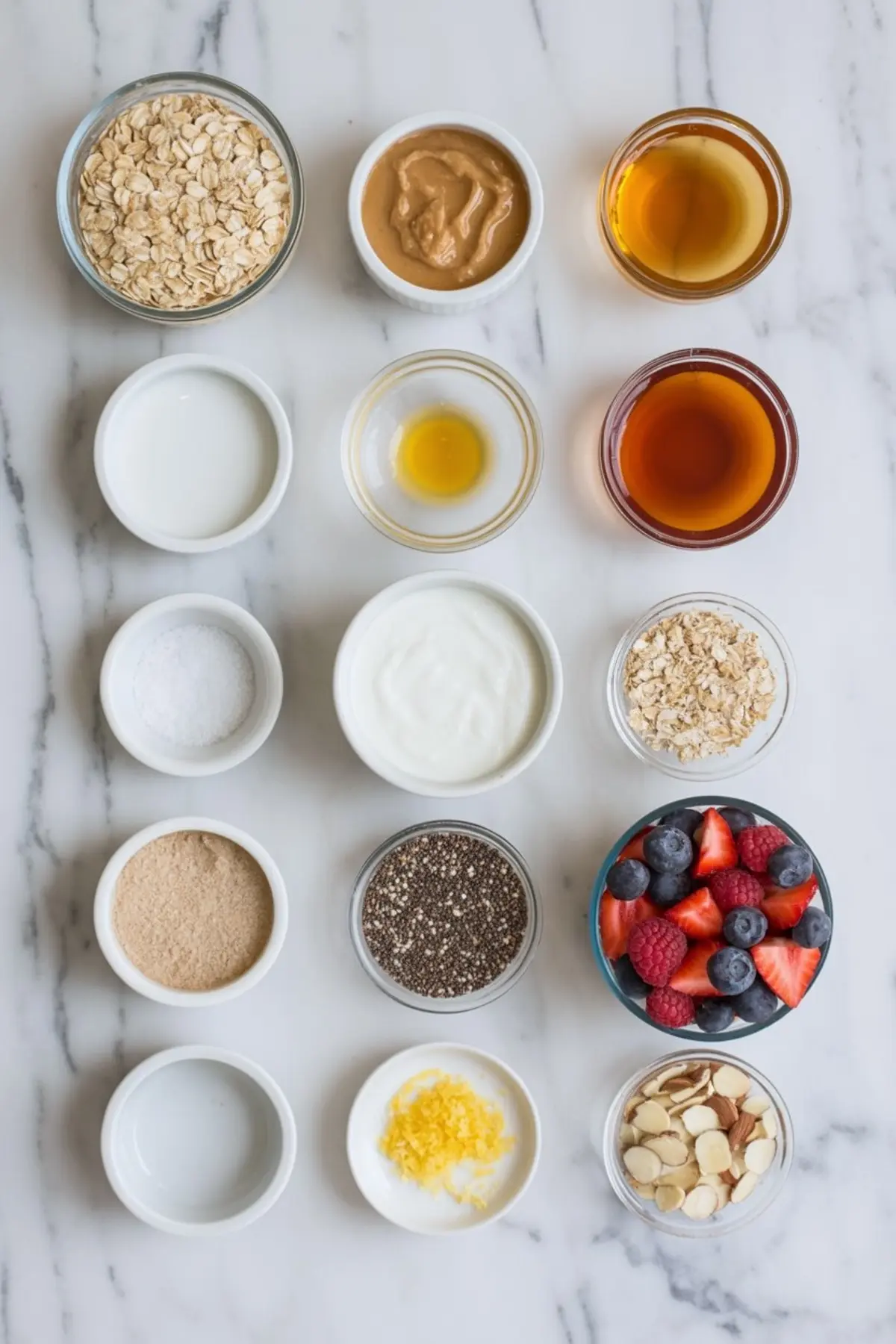 Flat lay of prepped ingredients for protein bars including oats, peanut butter, maple syrup, chia seeds, berries, yogurt, lemon zest, and protein powder in small bowls on a marble surface.
