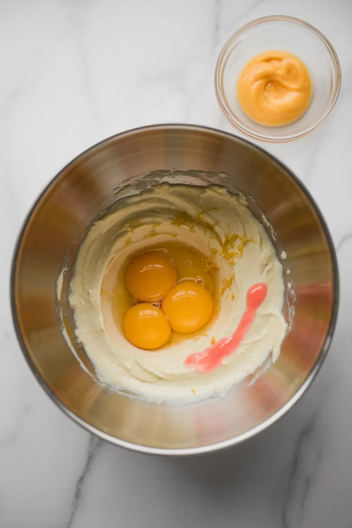 Metal mixing bowl with cake batter, showing egg yolks, blood orange zest, and a streak of vibrant blood orange juice, with a small bowl of orange curd nearby.

