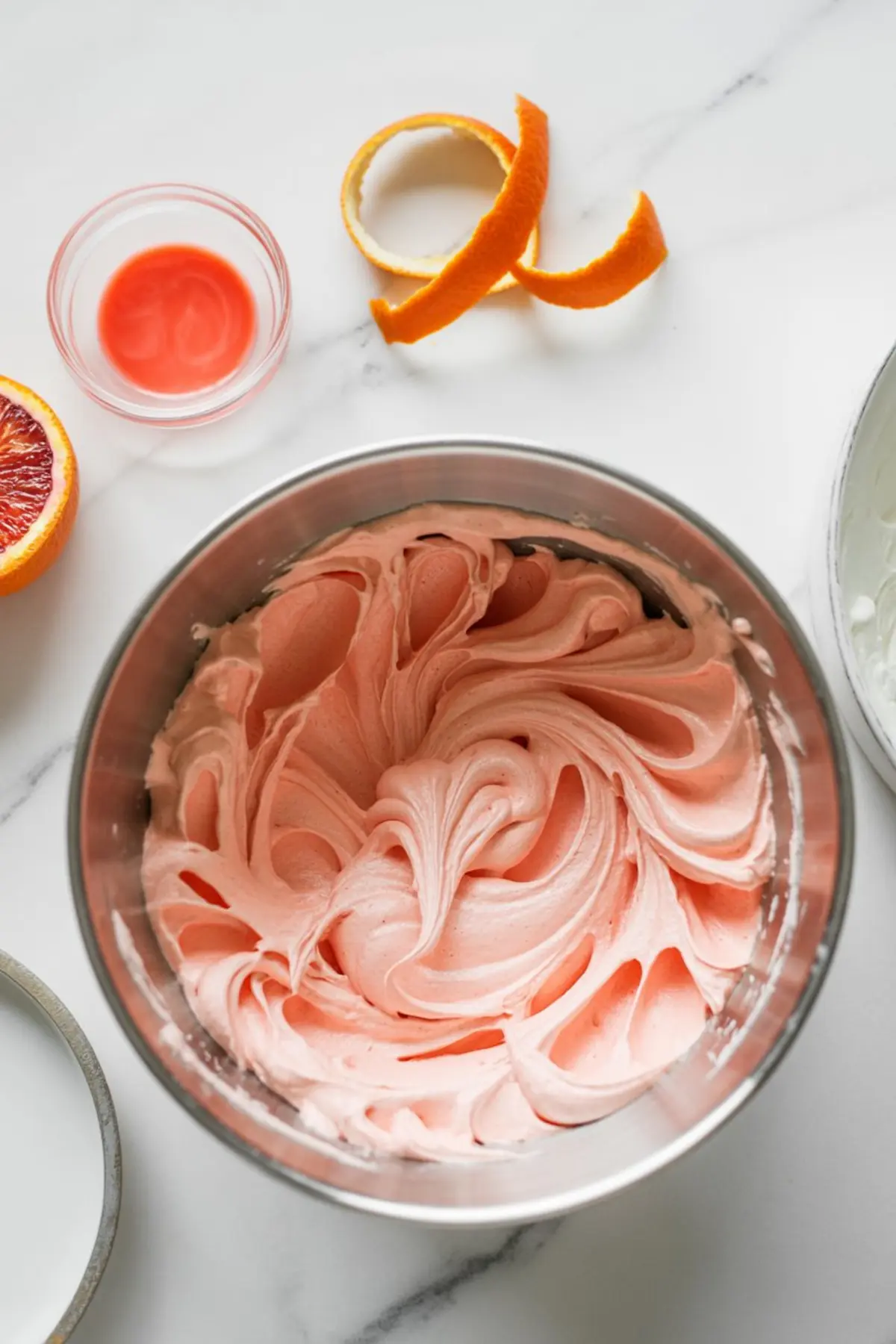 Pink whipped blood orange buttercream frosting in a mixing bowl, surrounded by orange peel curls and blood orange halves on a marble countertop.
