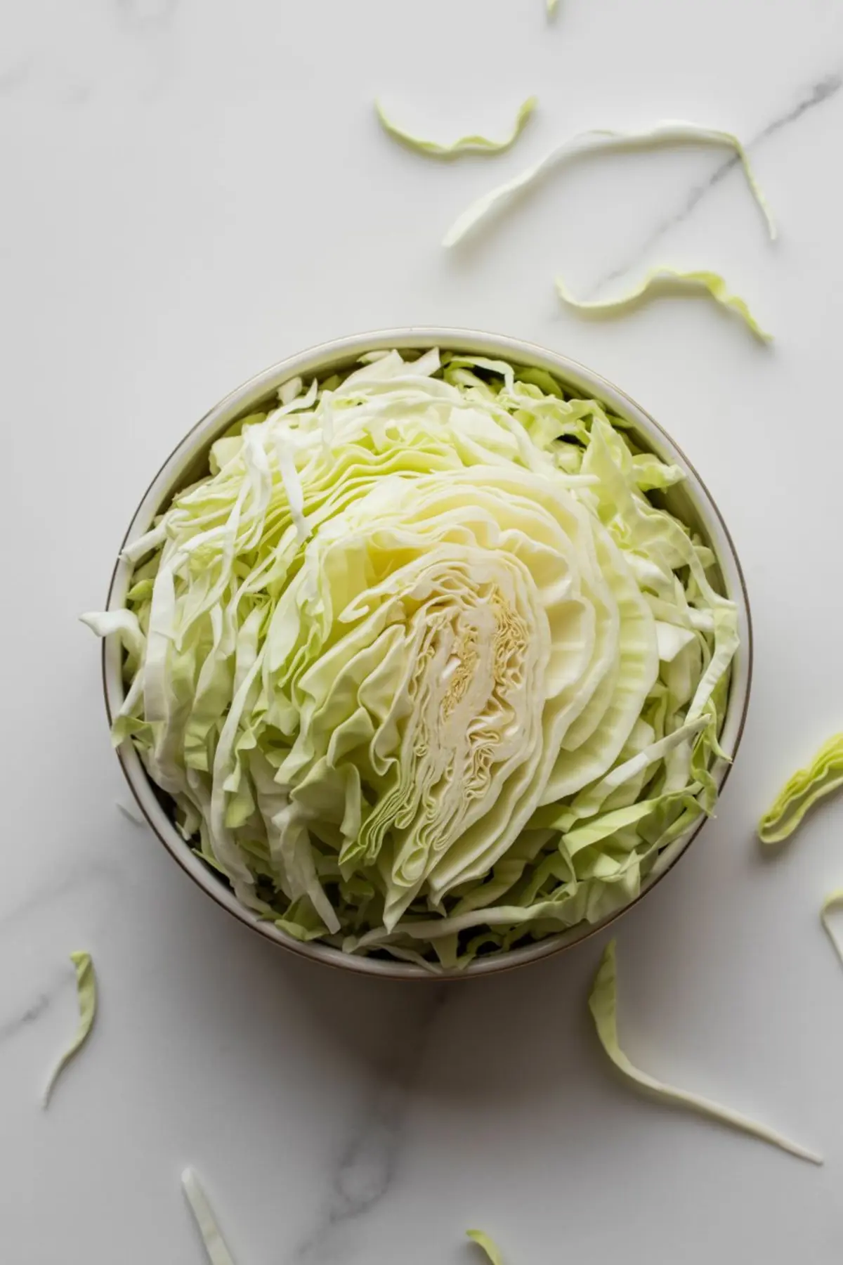 Freshly shredded green cabbage displayed in a round bowl, with loose strands scattered on a white marble surface, showcasing crisp texture and light green color.
