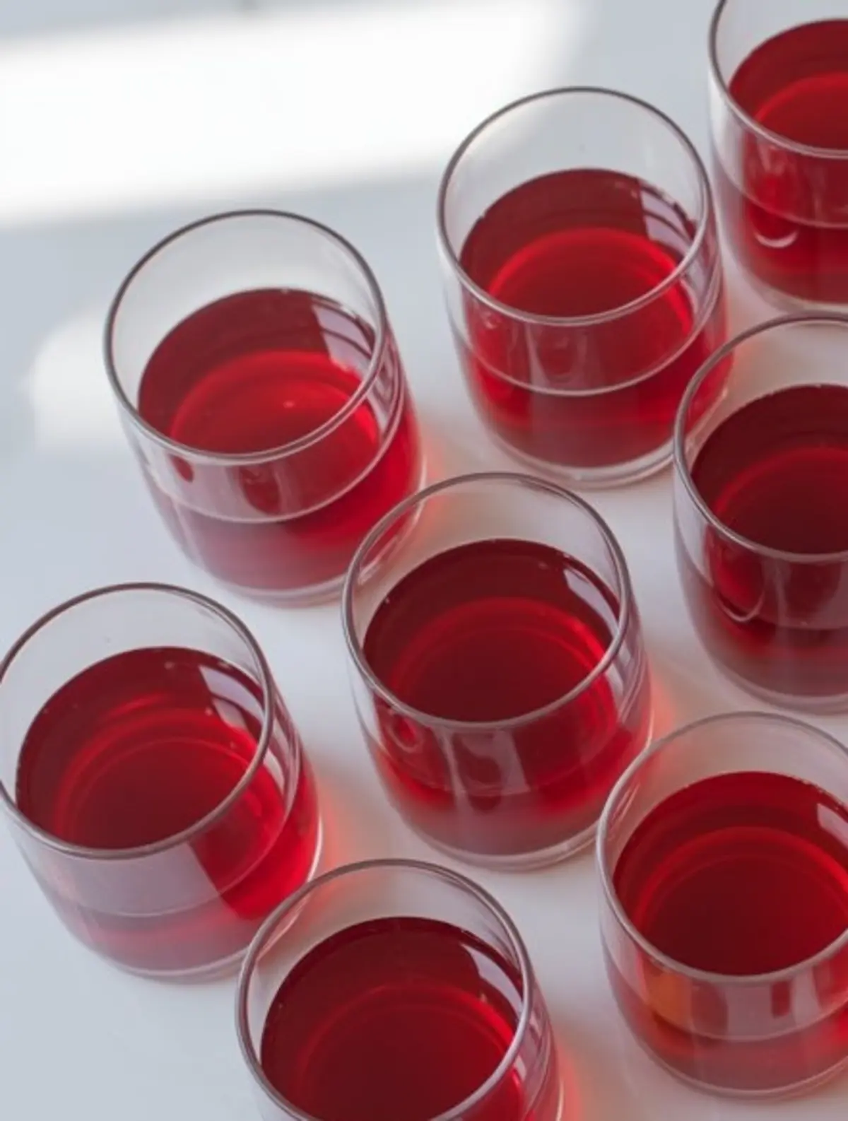 Clear glass cups filled with solidified red Jello arranged in a grid pattern on a white surface, capturing a top-down view with natural light reflections.