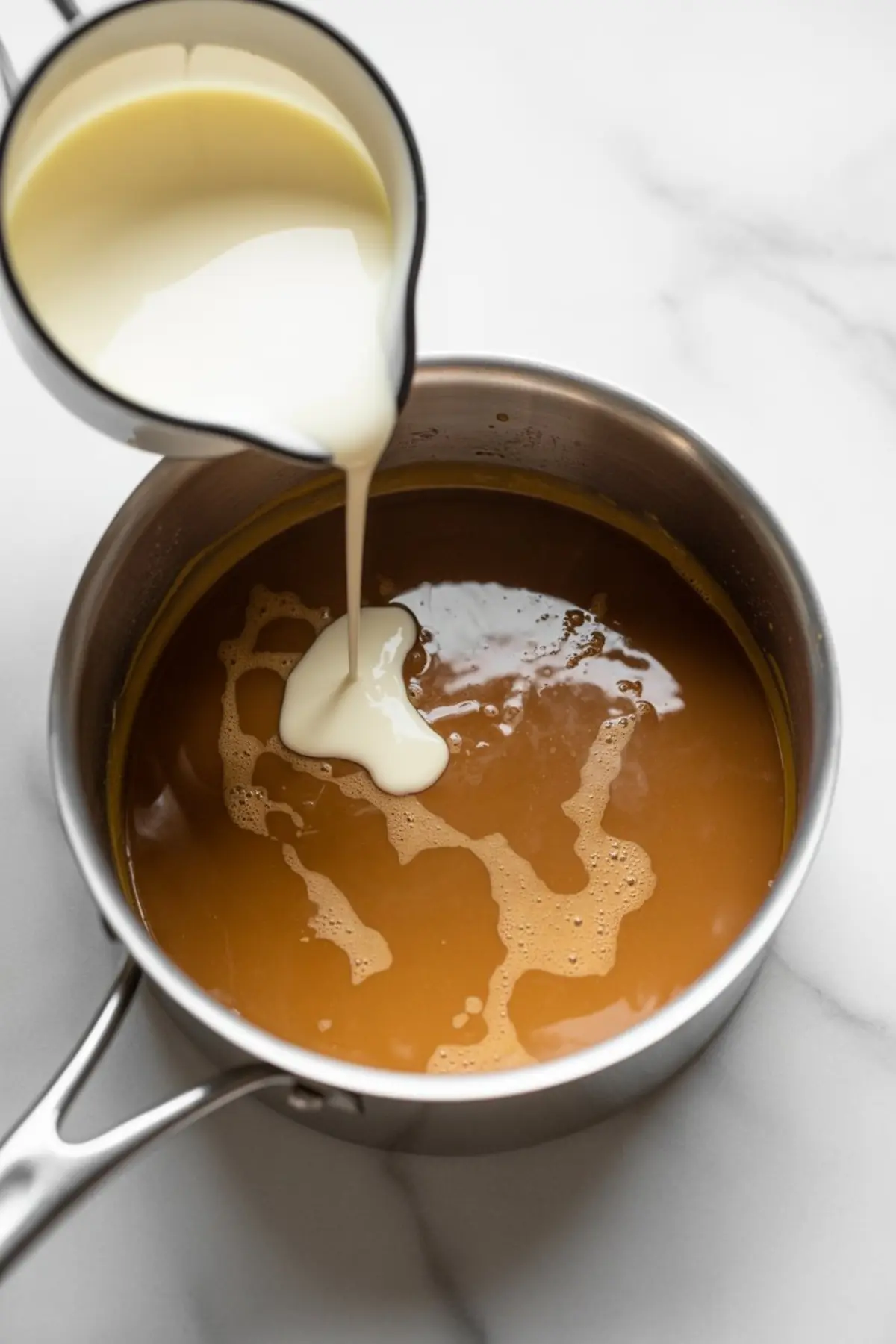 Heavy cream pours from a small pitcher into a saucepan of warm caramel sauce, showing the process of making homemade salted caramel for buttercream frosting.
