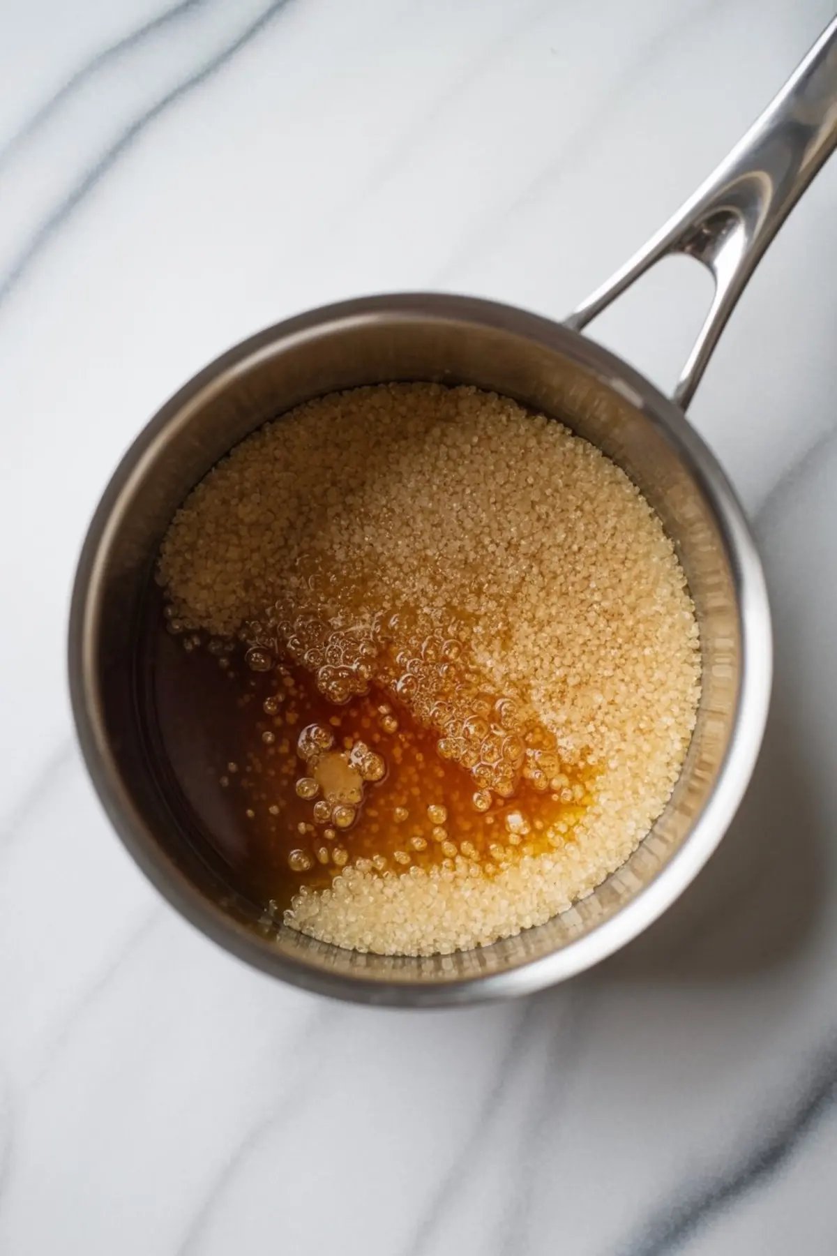 Granulated sugar melts and bubbles in a stainless steel saucepan, showing the early stage of caramel sauce cooking for salted caramel buttercream.
