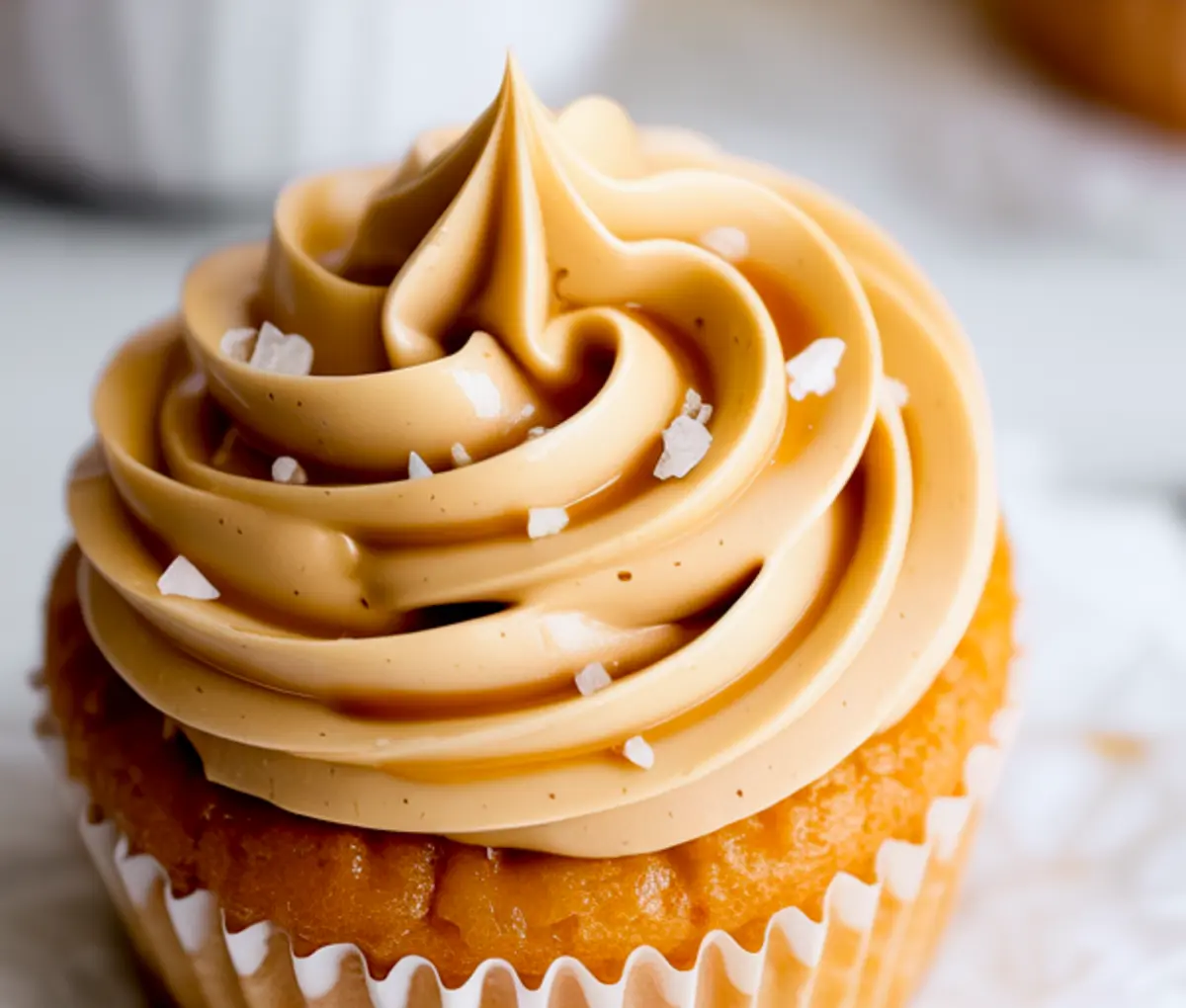 Vanilla cupcake sits in a paper liner topped with a tall swirl of salted caramel buttercream frosting and sprinkled with flaky sea salt.
