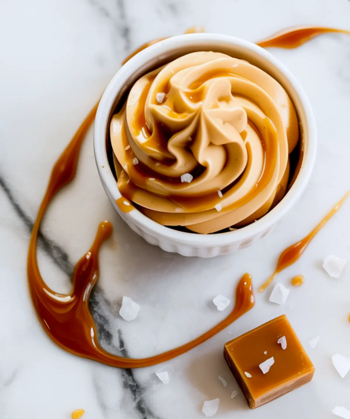 Bowl of salted caramel buttercream frosting sits on marble with caramel drizzle, flaky sea salt, and a caramel candy cube beside it.

